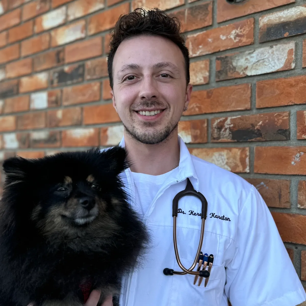 A smiling man in a white shirt with a stethoscope around his neck holding a black and tan small dog, standing in front of a brick wall.