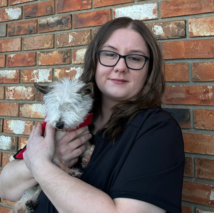 A woman with glasses holding a small, fluffy dog with white and black fur in front of a brick wall.