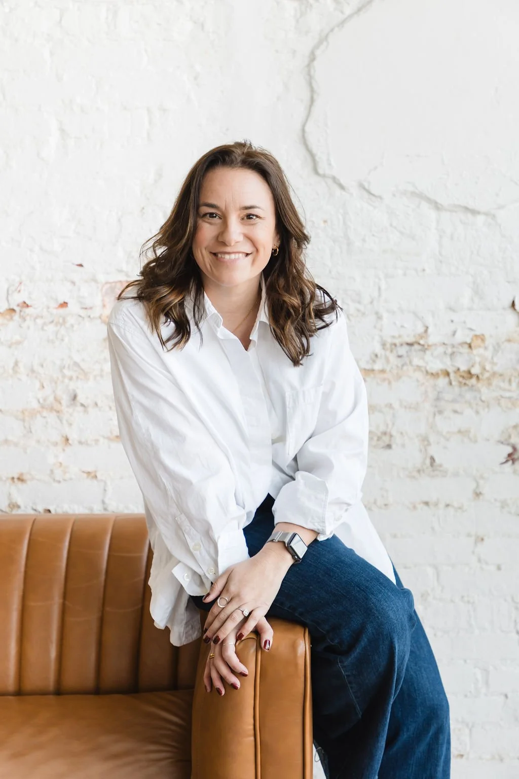 A woman with brown wavy hair smiling while sitting on a tan leather sofa against a white brick wall, dressed in a white button-up shirt and dark jeans.