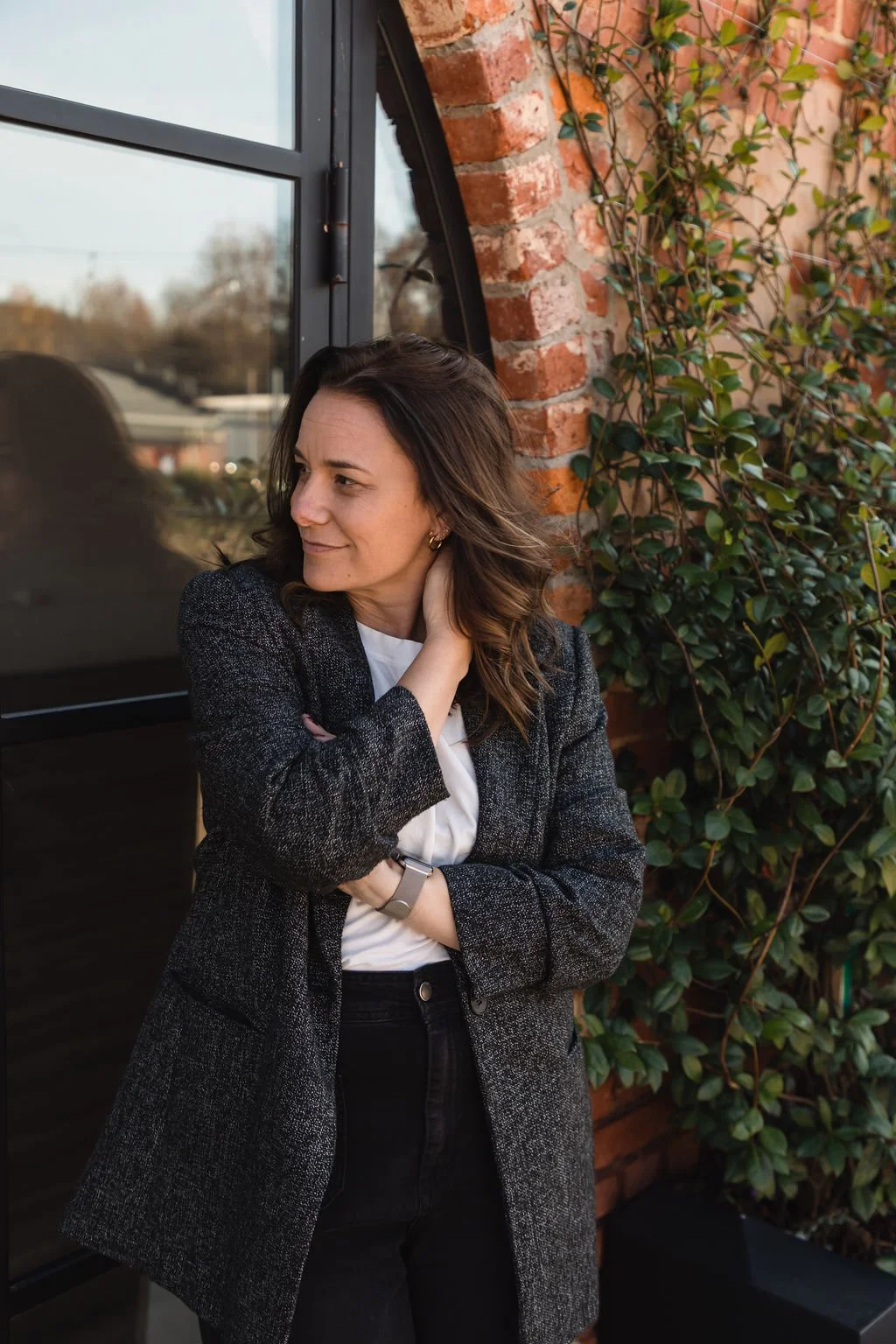 A woman with brown hair wearing a black blazer and white shirt standing outdoors next to a brick wall and green leafy vine, looking to her left.