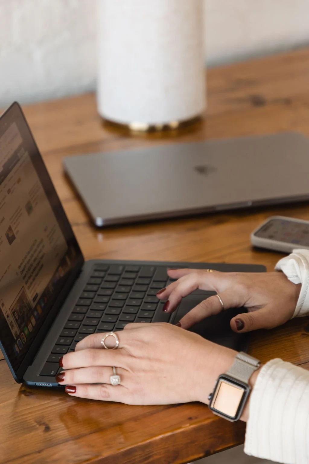 Person using a laptop on a wooden table, with a smartphone and a closed laptop nearby, and a white lamp in the background.