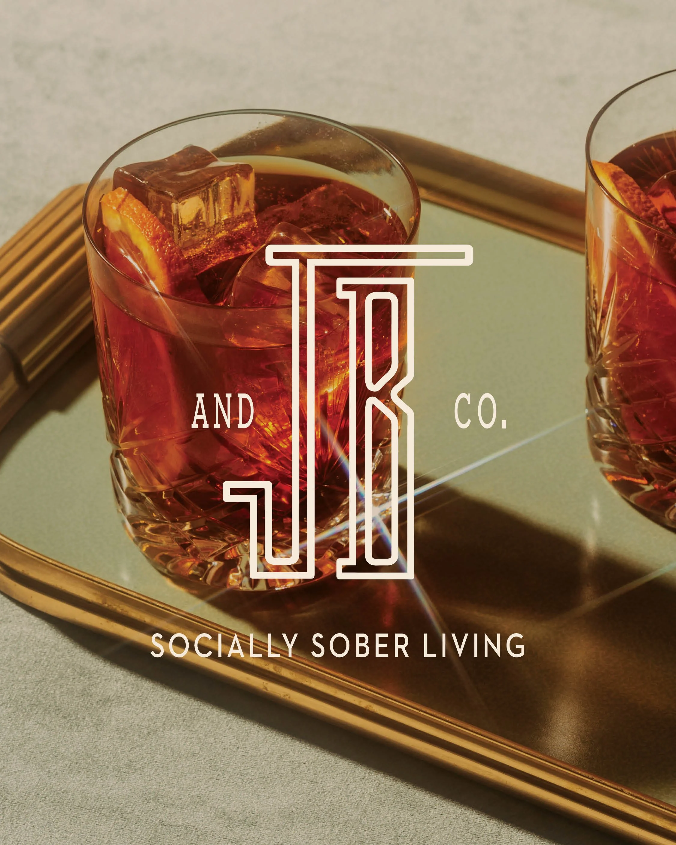 Two glasses of iced beverages with orange slices on a gold tray, overlaid with the JB & Co logo and text for 'Socially Sober Living'.