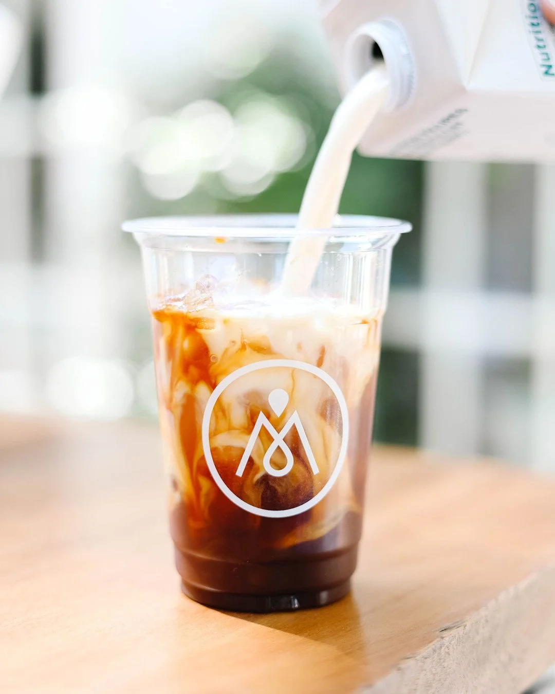 Maisonette branding example of coffee being poured into a plastic cup with ice with logo on the cup, on a wooden surface, blurred background.