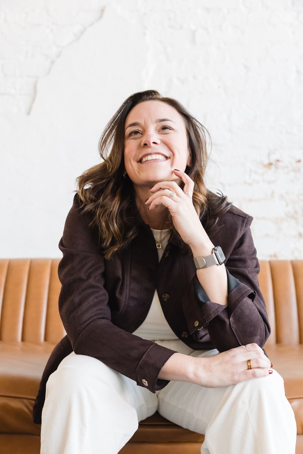A woman with brown hair smiles while sitting on a tan couch against a white wall with exposed brick.