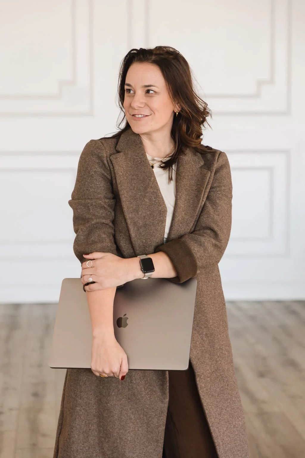 A woman with short brown hair, wearing a brown coat with rolled-up sleeves, stands indoors holding a closed silver Apple MacBook laptop and smiling to the side.