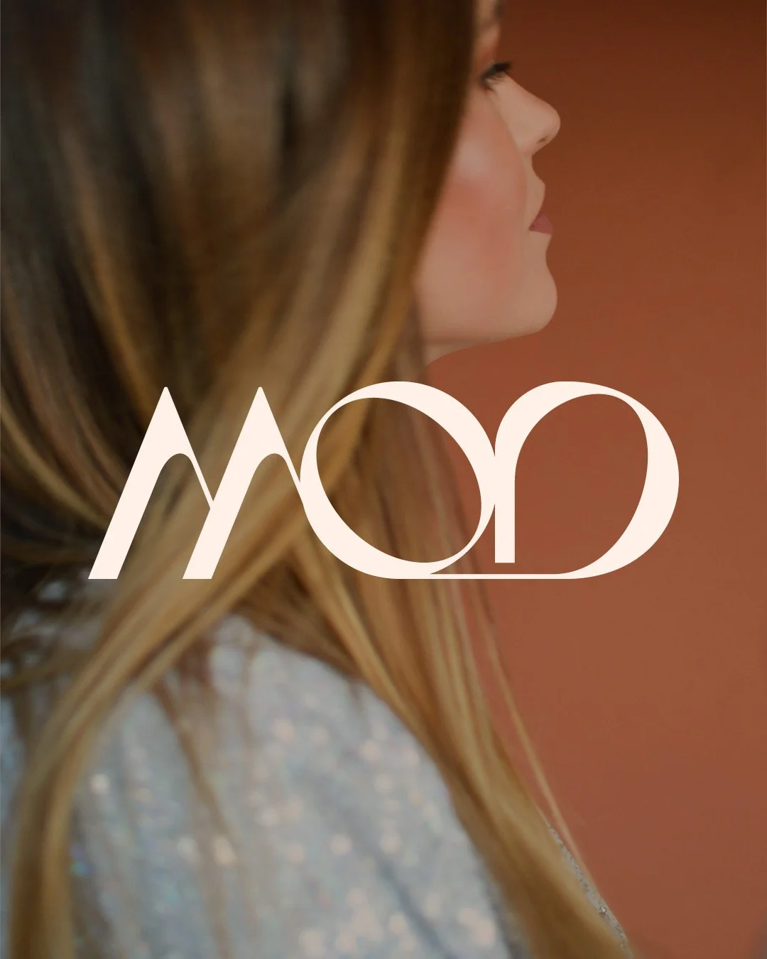 Close-up profile of a woman's face with long, wavy brown hair and a sparkly top, with a blurred warm background and a logo variation for the Mod Shop.