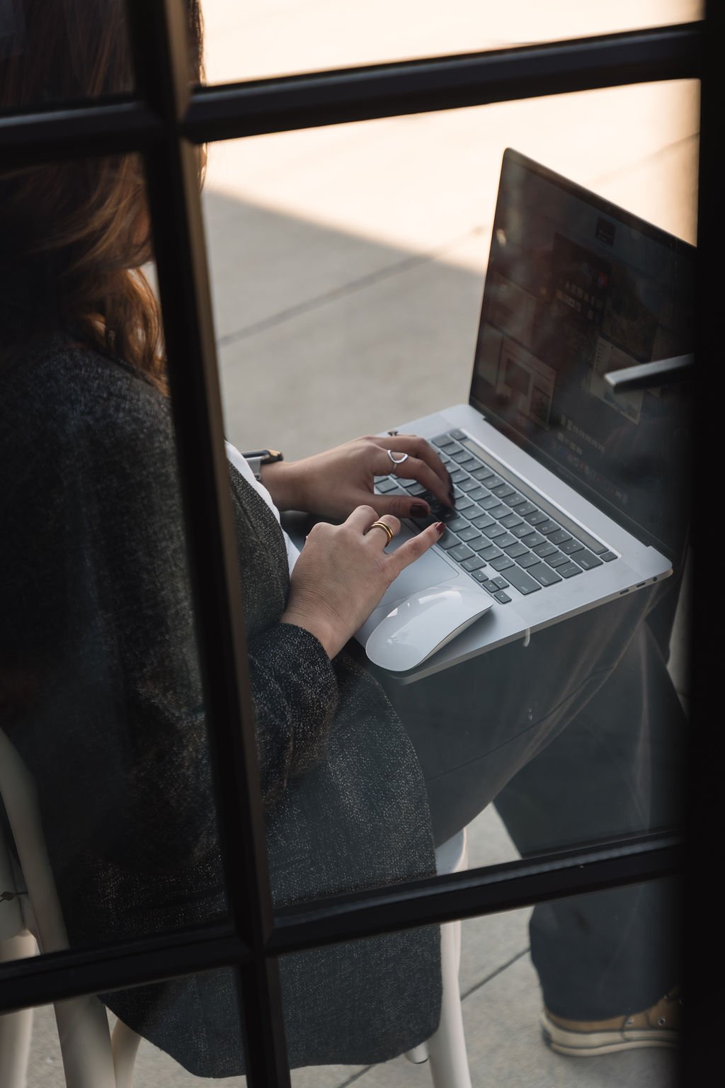 A person using a laptop at a desk, viewed through a window with black grid framing.