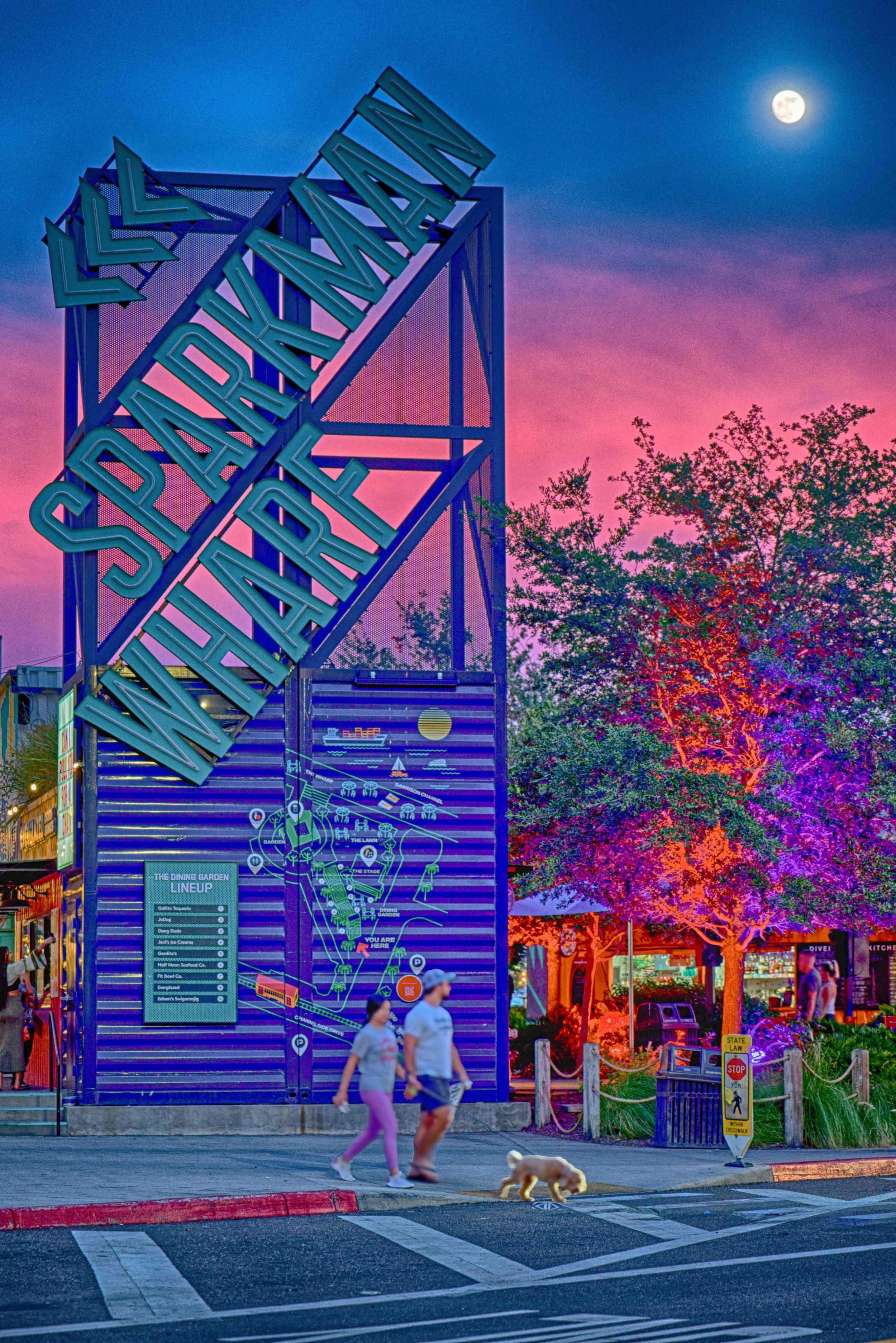 People walking a dog in front of a vibrant, illuminated sign for Sparkman's Wharf at dusk, with a colorful sunset sky, a full moon, and a lit purple tree.