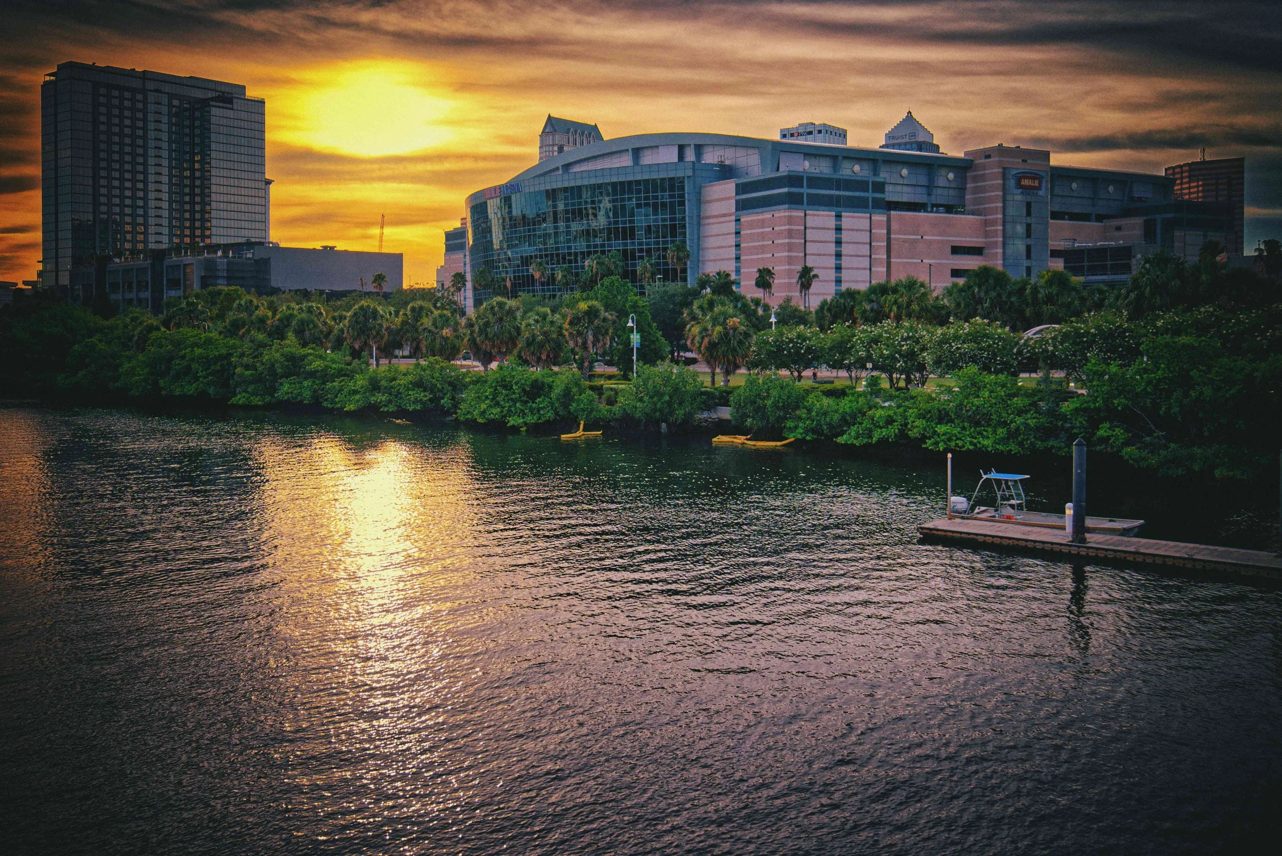 Sunset over a cityscape with modern buildings, palm trees, and a body of water with a small dock in the foreground.