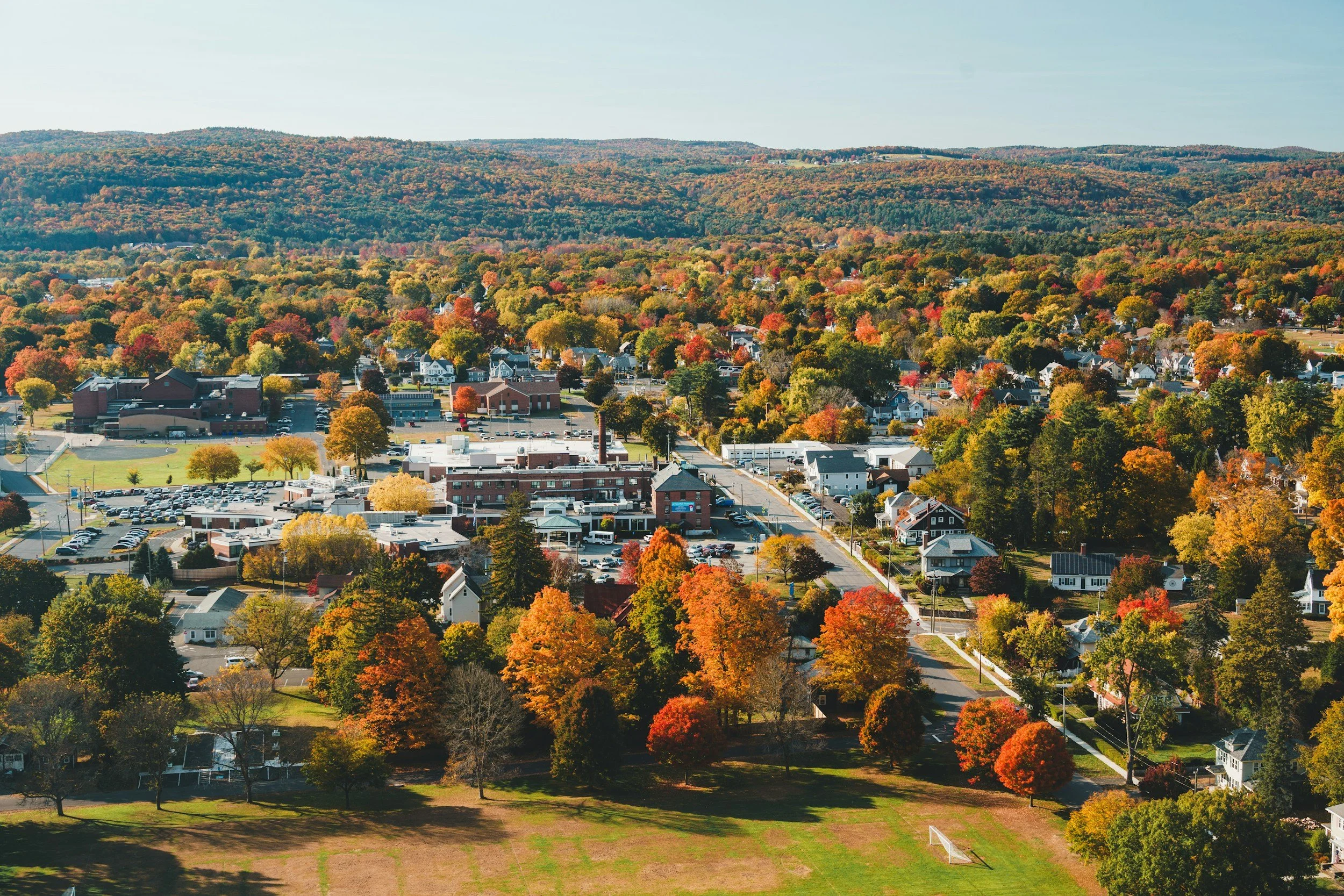 Aerial view of a town with colorful autumn trees, residential houses, a school, and a hill in the background on a clear day.