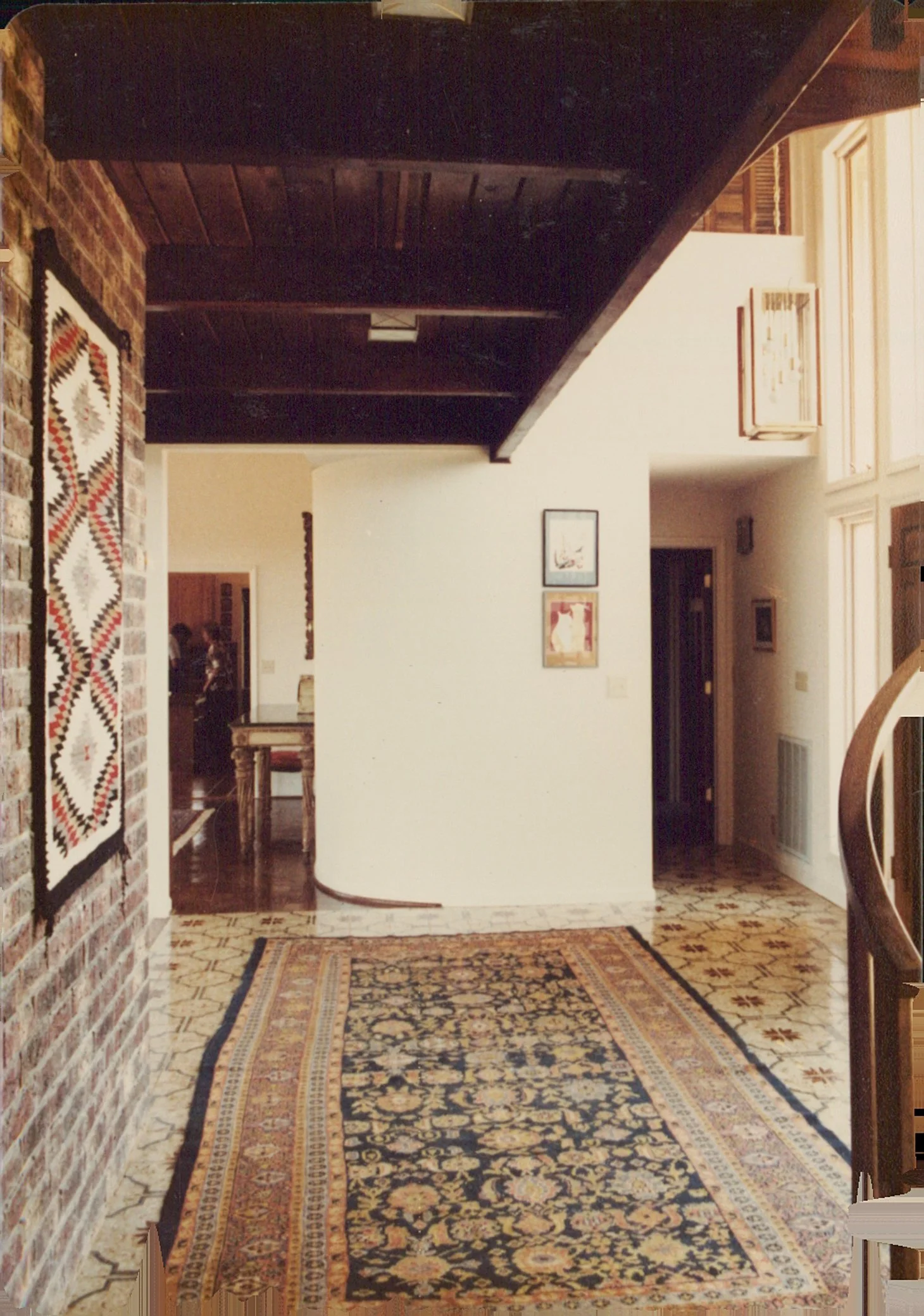 Interior of a home featuring a patterned rug on the floor, a brick wall on the left with a decorative textile, a wooden ceiling, and framed artwork on white walls.