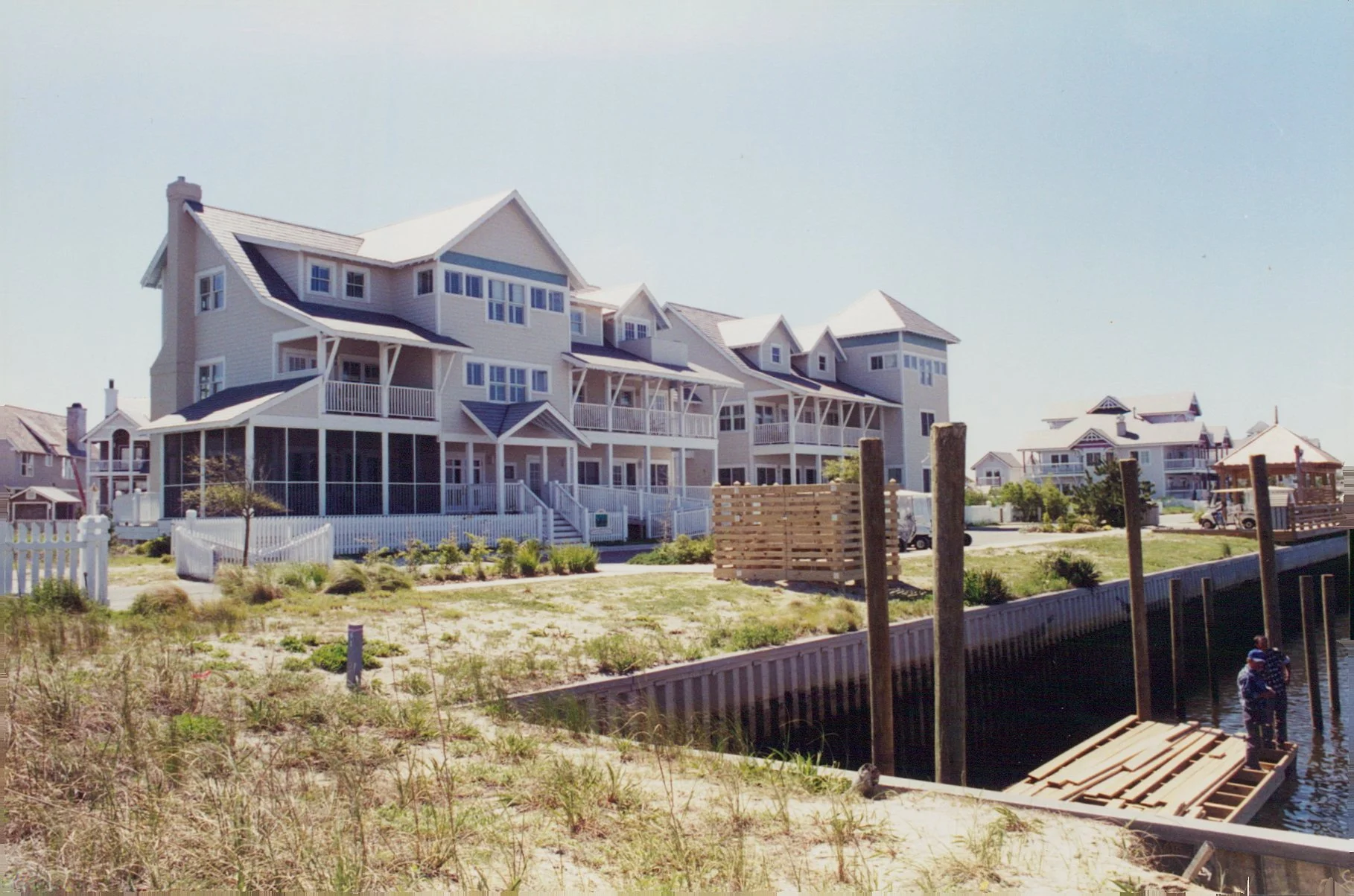 A large multi-story beach house with multiple decks and white fencing, located near a canal with a dock and a few people standing on the dock.