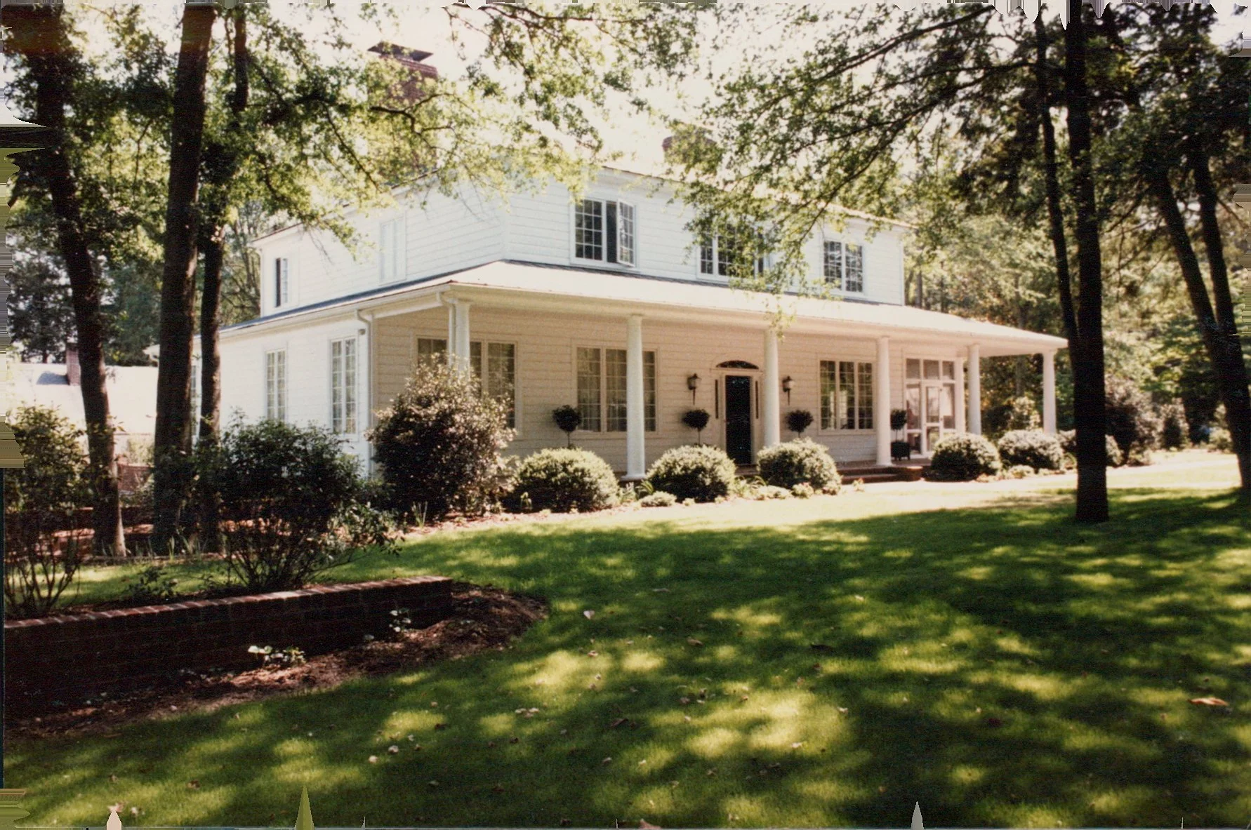 A large white house in the Pinehurst Village designed by Jose Camina with a wrap-around porch, surrounded by trees and a well-maintained lawn with bushes.