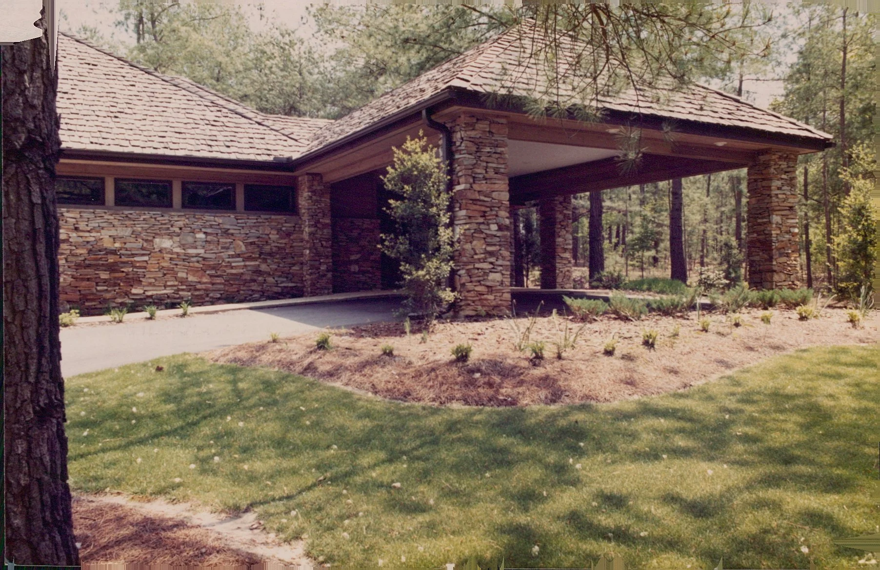 Modern house with stone walls, surrounded by trees and a manicured lawn, featuring a covered porch area.