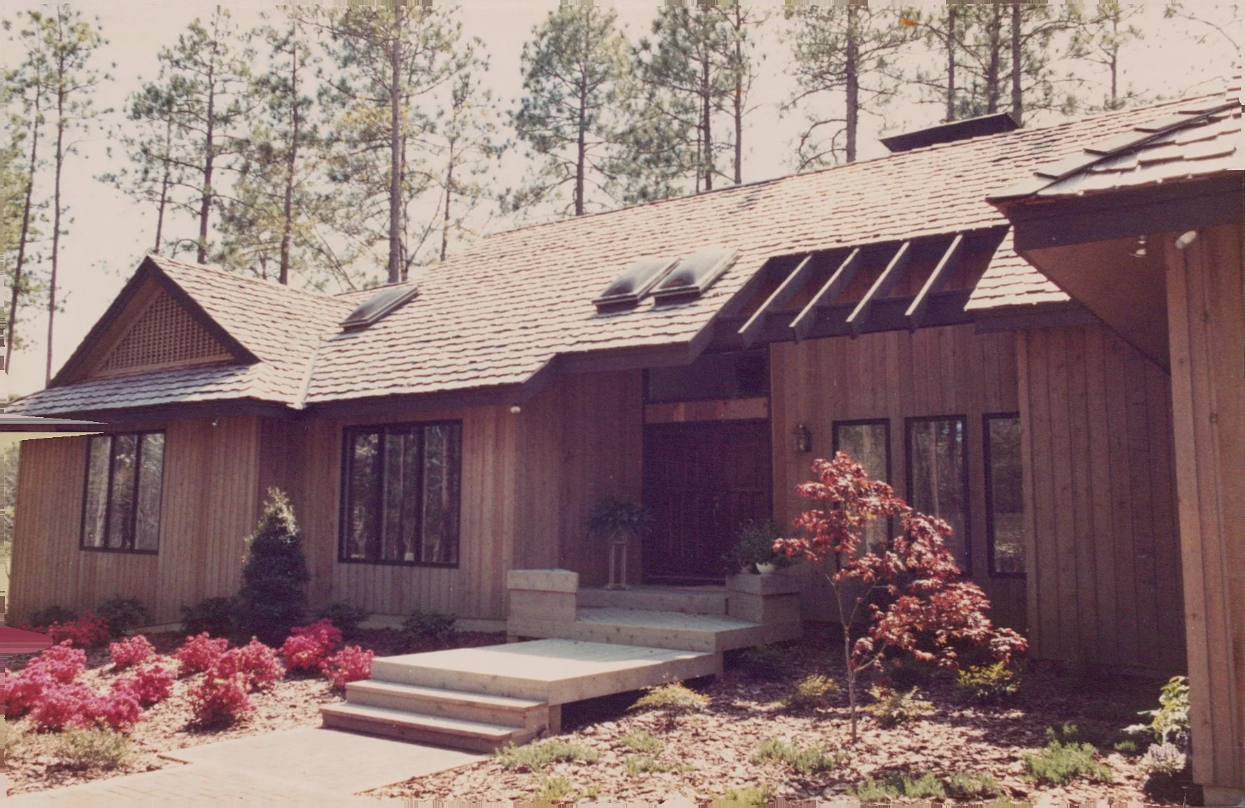 A wooden house with a sloped roof, skylights, large windows, and a front porch with steps, surrounded by landscaped garden with flowers and small trees.