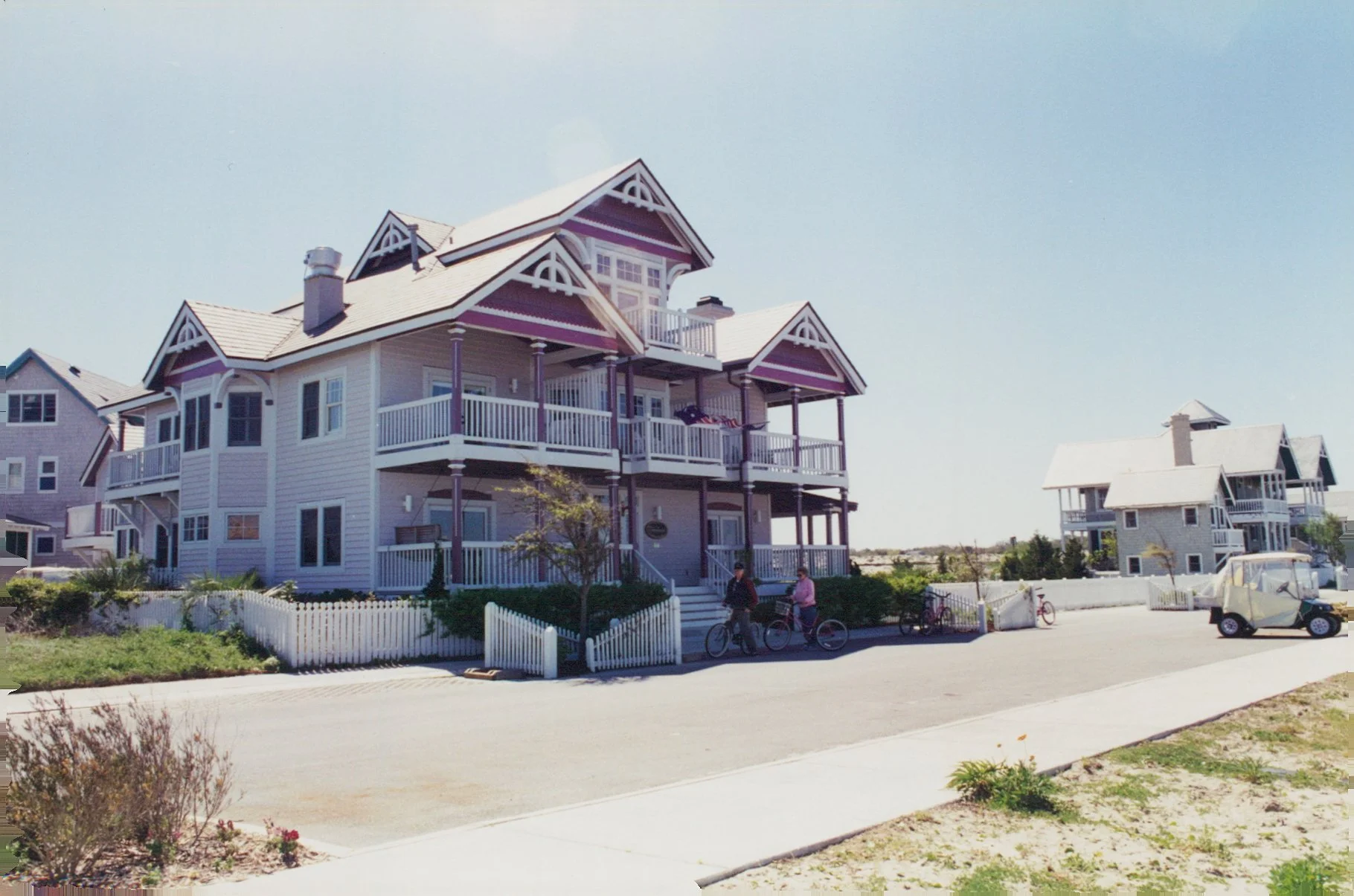 A large, white, three-story beach house with purple accents and a wraparound porch, located in a coastal area. Several people with bicycles are on the sidewalk in front of the house, and other similar houses are visible further down the street. The sky is clear and sunny.
