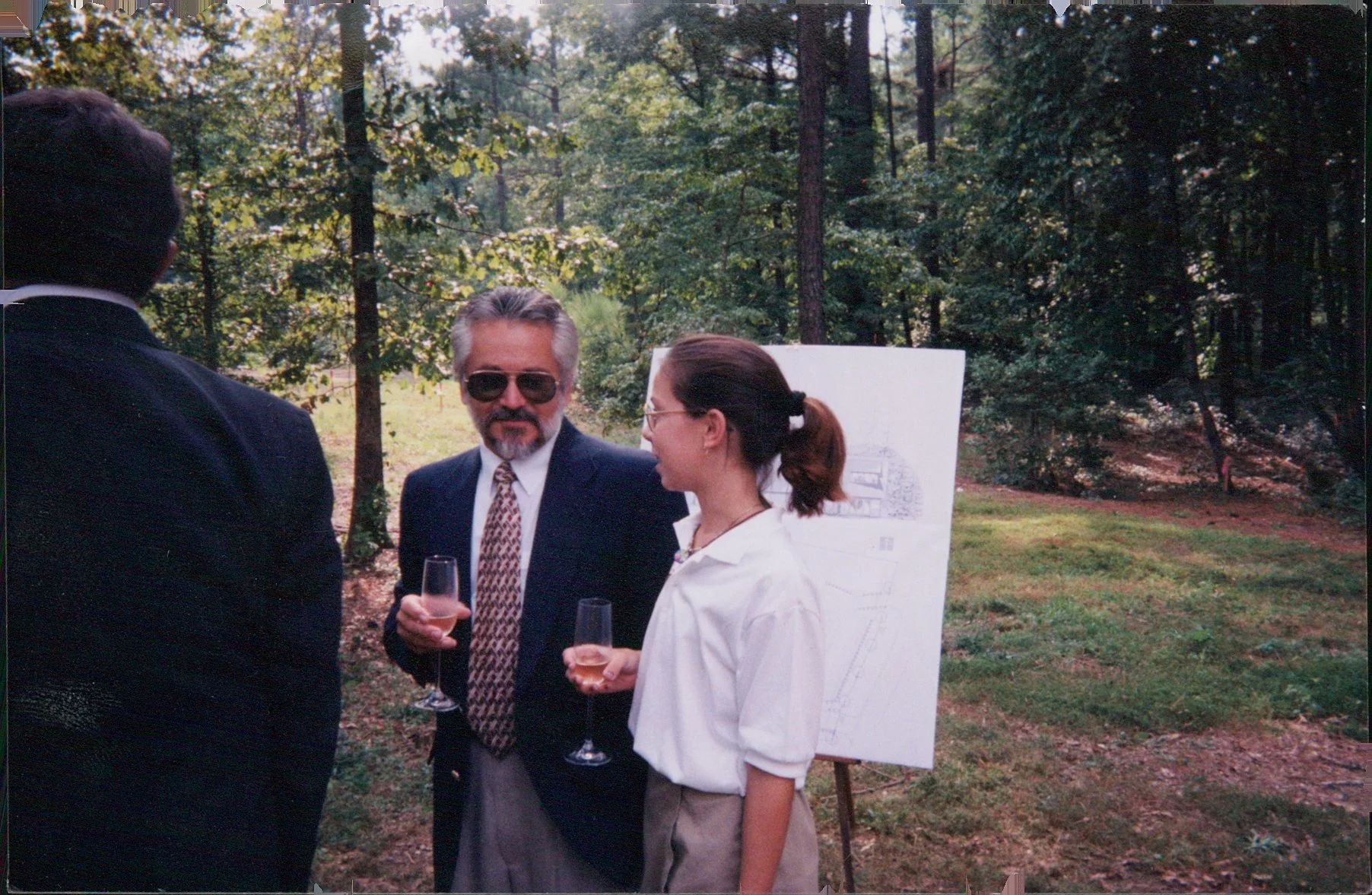 Three people having a conversation outdoors in a wooded area, each holding a glass of rosé wine, with a whiteboard displaying a sketch or plan behind them.