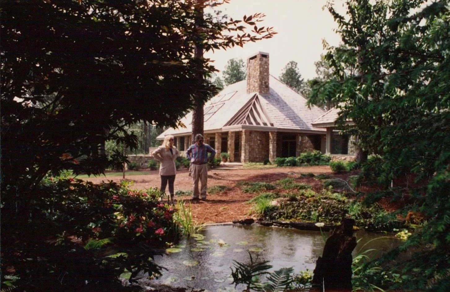 A couple standing outdoors near a pond in front of a brick house under construction, surrounded by greenery and trees.
