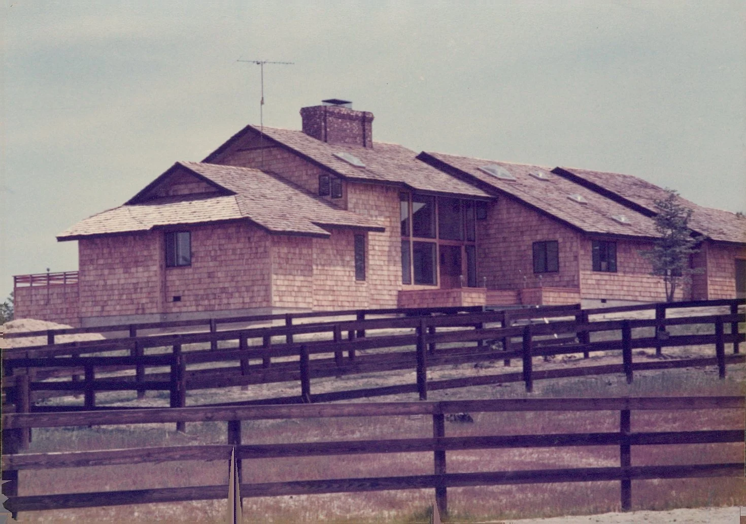 A large house made of reddish-brown wooden shingles with multiple levels and rooflines. It has several windows, a brick chimney, and a central glass section. A black wooden fence surrounds the house, and there is a small tree to the right, with a partly cloudy sky overhead.
