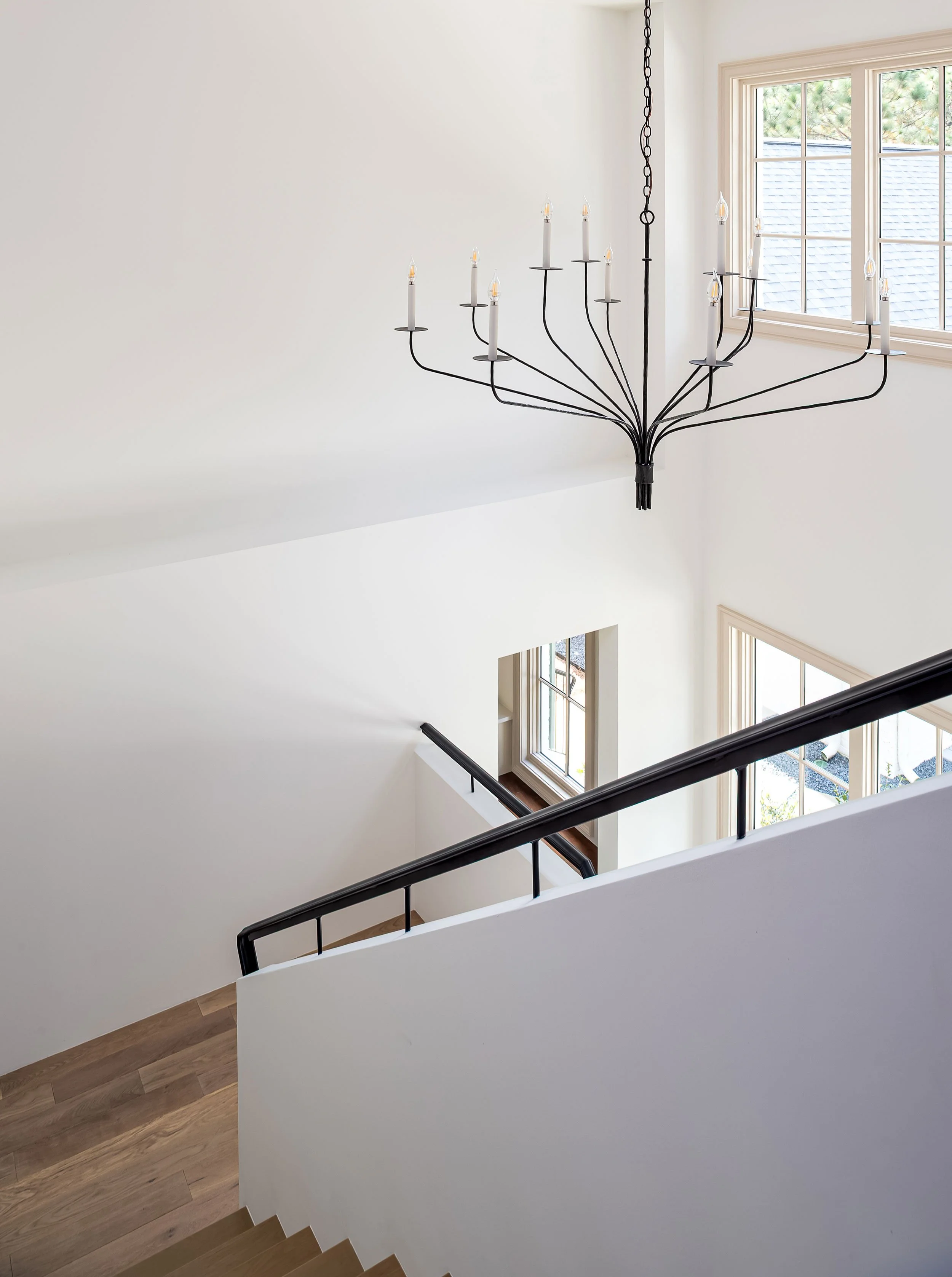 Interior view of a staircase with black handrail and wooden steps, white walls, large windows, and a black chandelier with candle-like lights hanging from the ceiling.