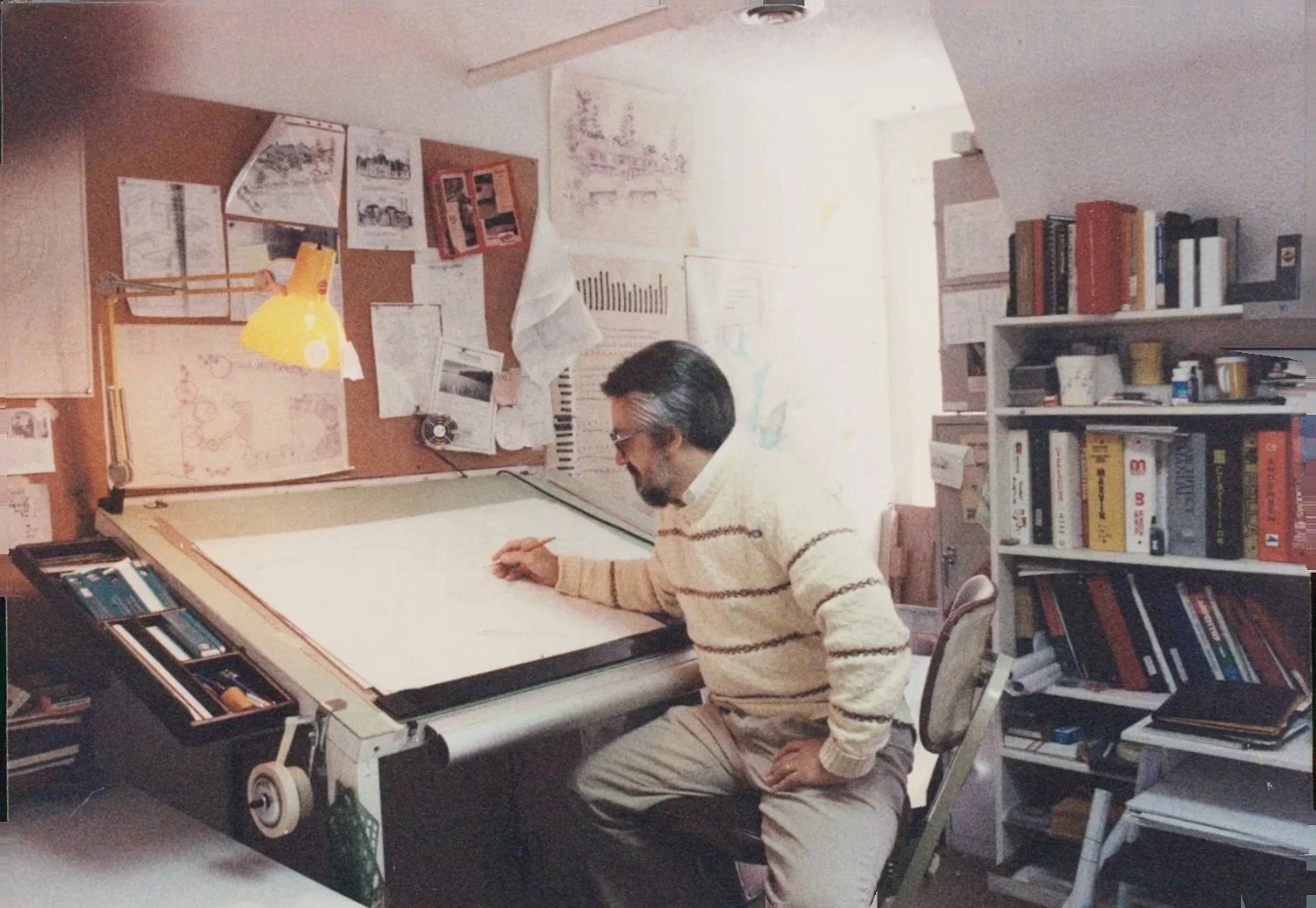 A man sitting at a drafting table in a cluttered office, drawing on large paper, with shelves of books and a bulletin board behind him.