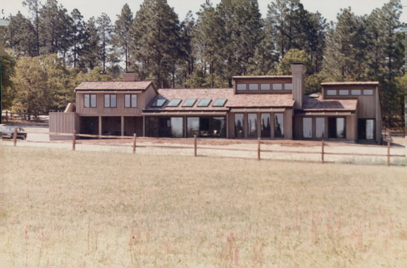 A modern architectural forward home built in the 1980s featuring atwo-story house made of wood with large windows, located in a wooded area with tall trees in the background and a grassy field in front, surrounded by a wooden fence.