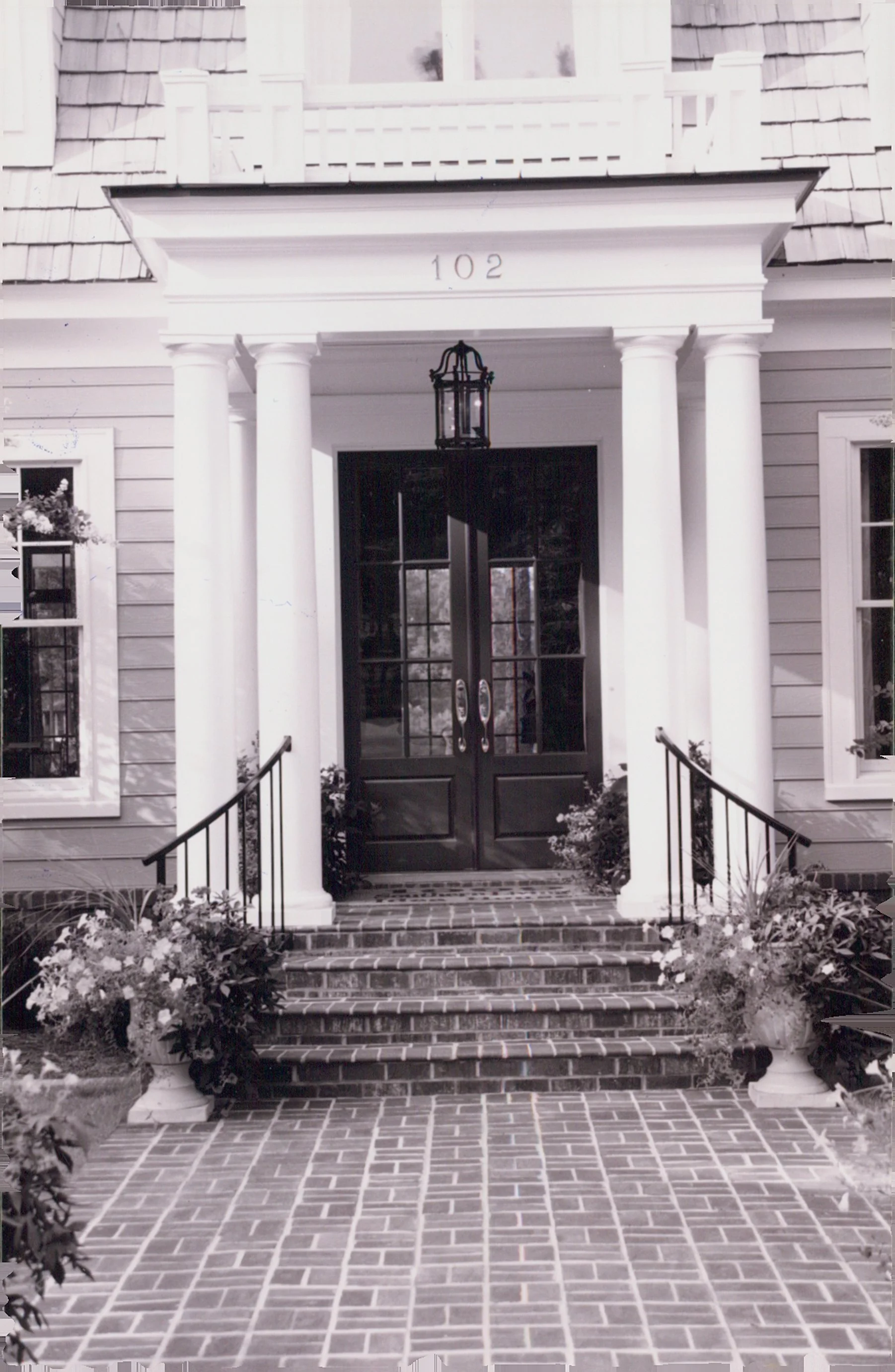 Front entrance of a house with a brick pathway, steps, black double doors, white columns, potted plants, and a lantern hanging above the door.