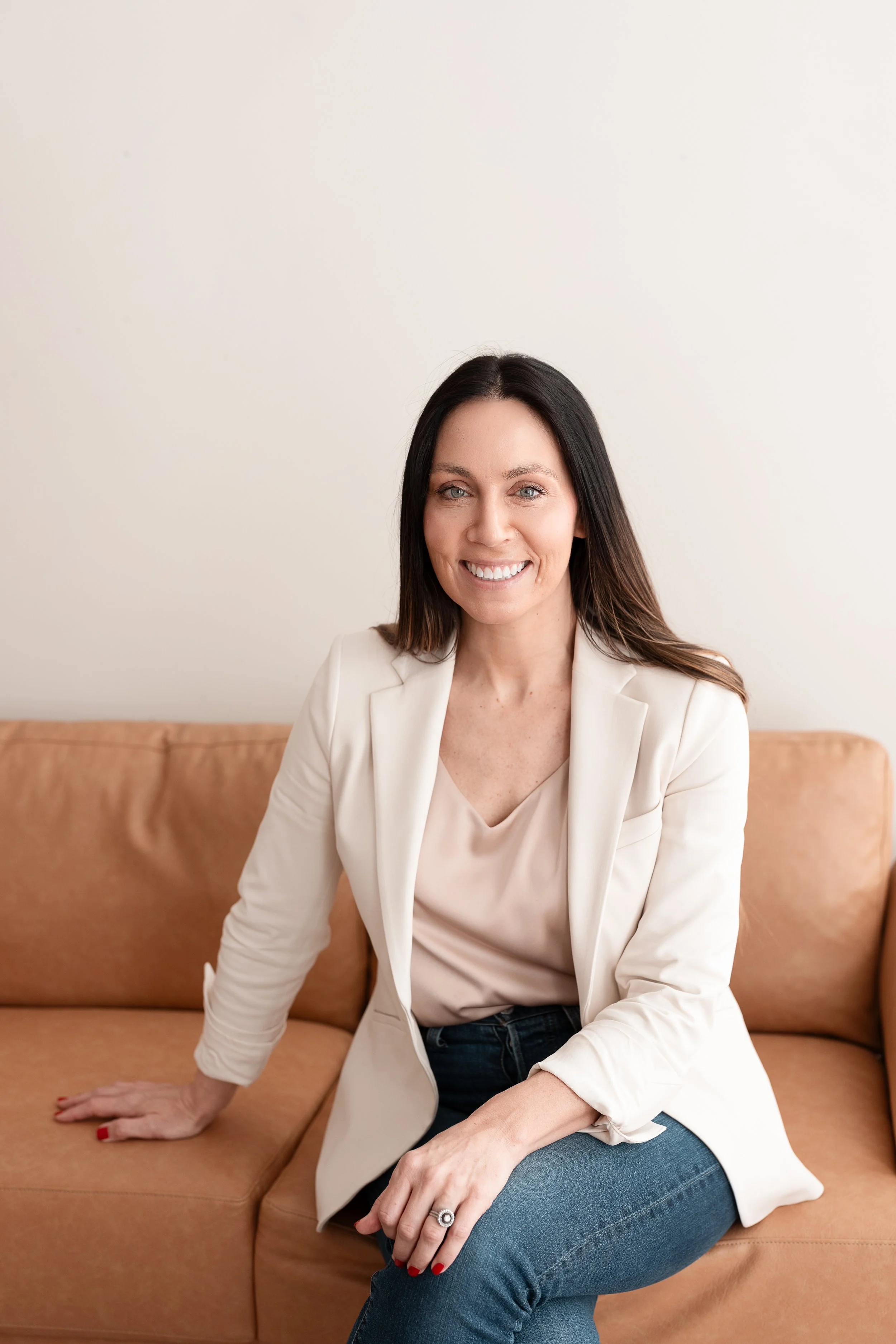 A woman with medium-length dark hair, smiling, wearing a white blazer, light pink blouse, and blue jeans, seated on a tan sofa against a plain off-white wall.