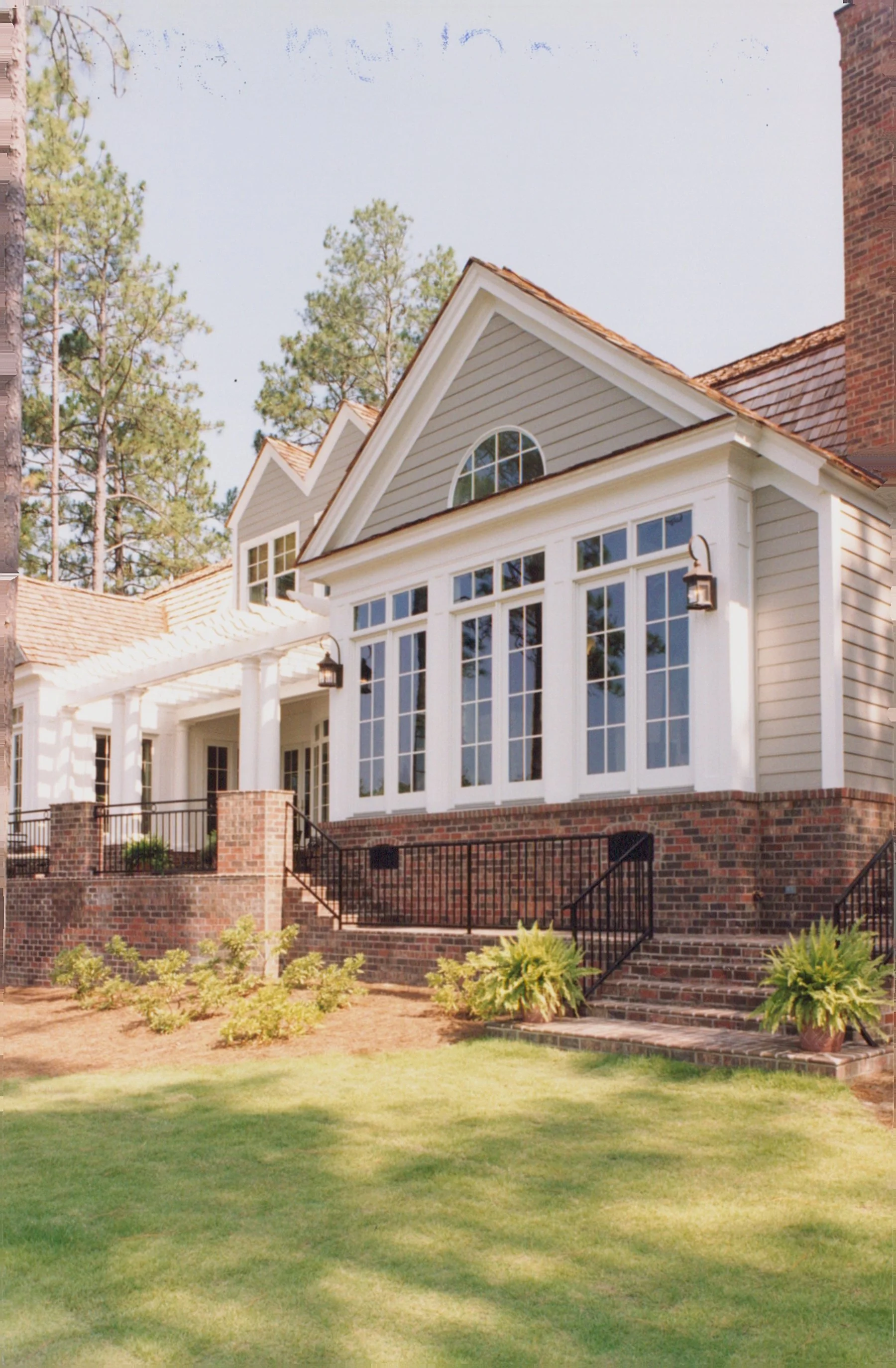 A large house with a brick foundation and white siding, featuring expansive windows and a porch with columns. The house is surrounded by a well-maintained lawn and some small bushes, and tall trees are visible in the background.