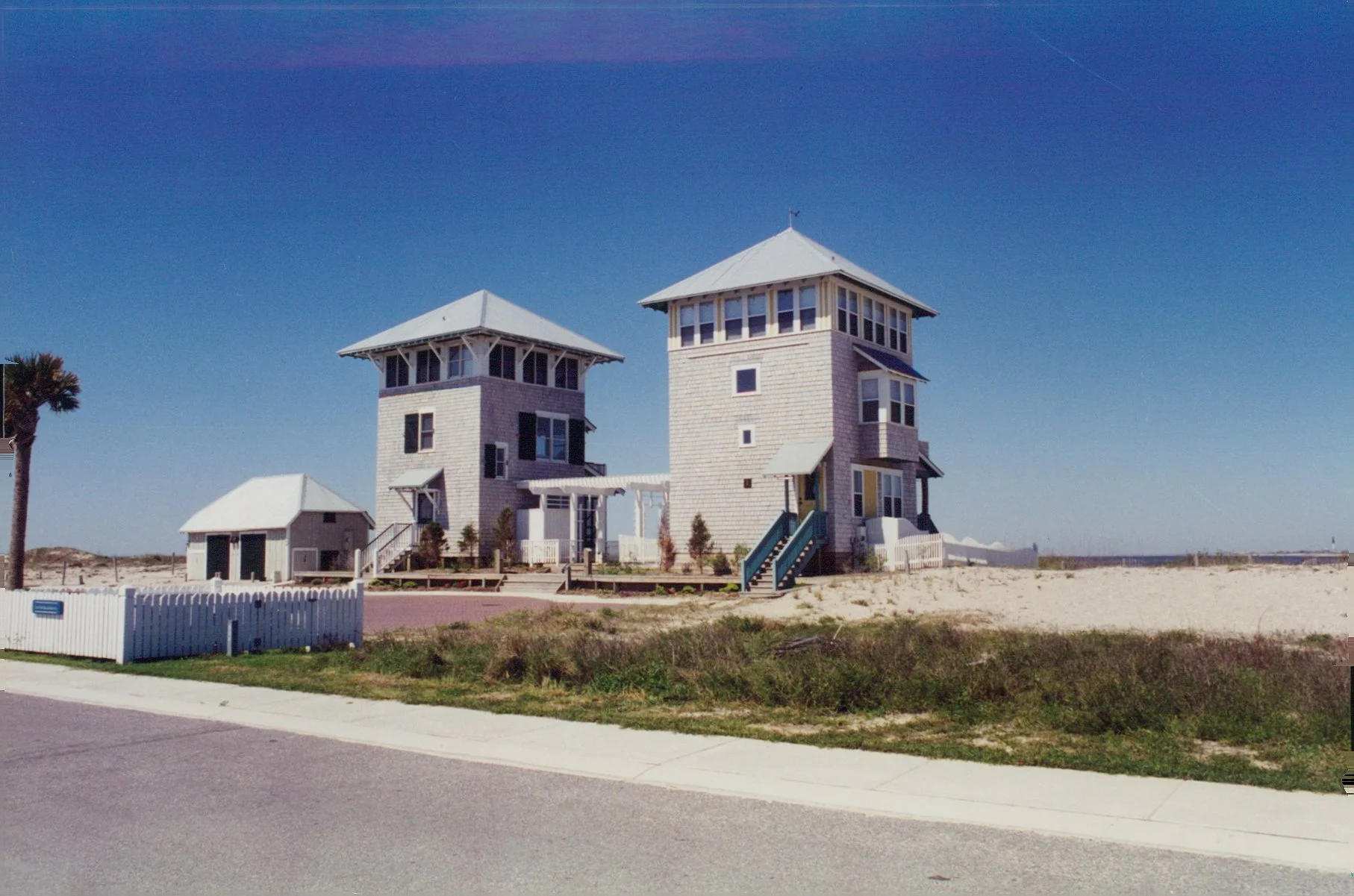 Two white houses with multiple windows and stairs, situated on a sandy beach with a palm tree and a white picket fence in the foreground, under a clear blue sky.