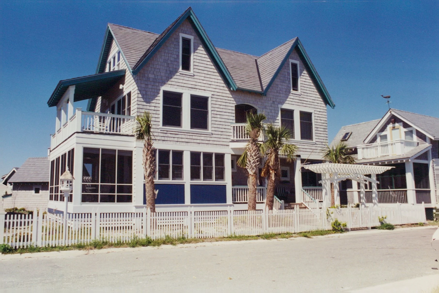 A multi-story beach house with a light gray exterior, dark teal trim, and multiple gabled roofs. The house features large windows, a wrap-around porch, and a small balcony. Palm trees and a white picket fence surround the house under a clear blue sky.