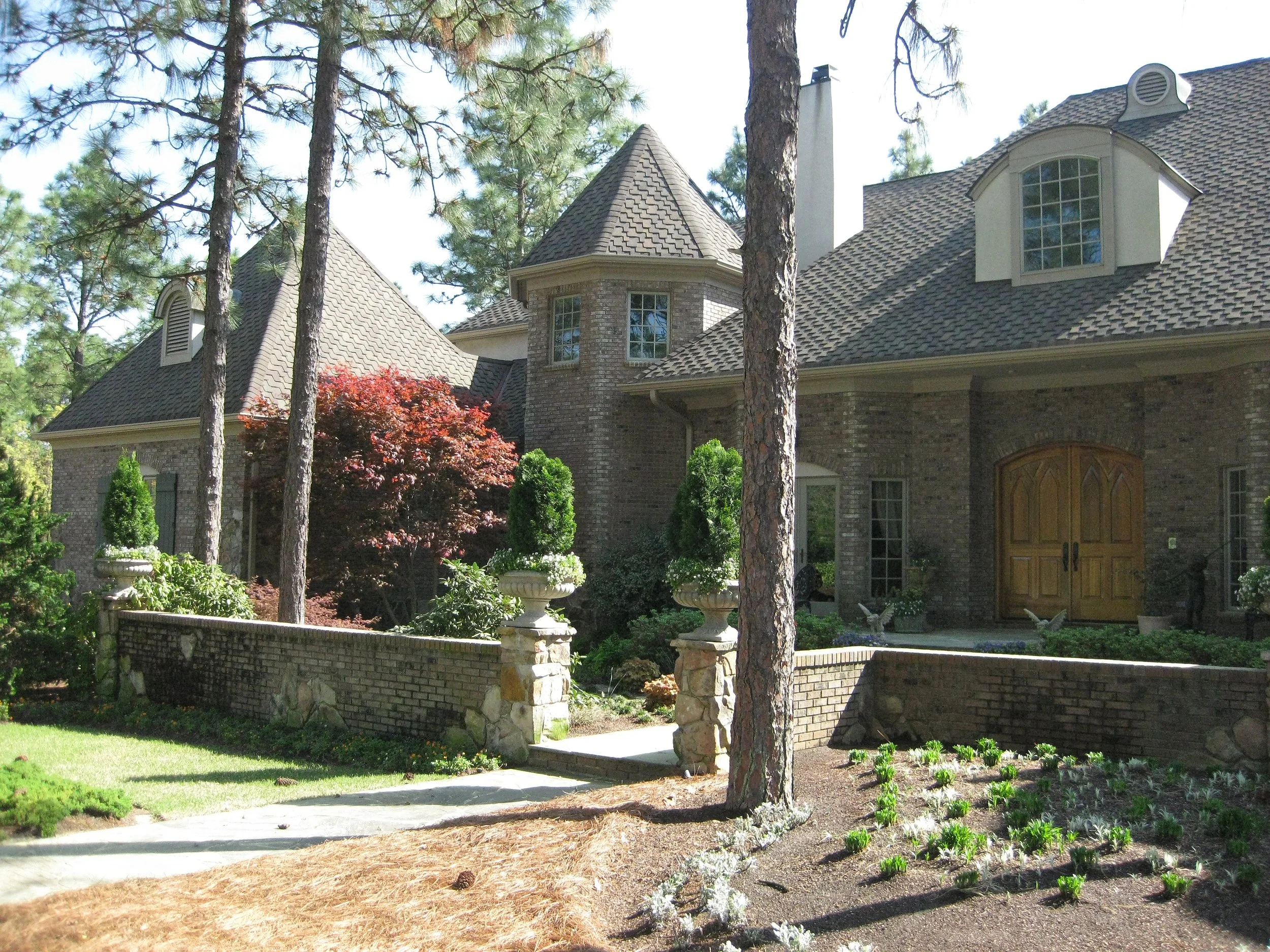 A large brick house with a gabled roof and multiple windows, surrounded by tall trees, a stone and brick fence, and a landscaped garden with shrubs and flowering plants.