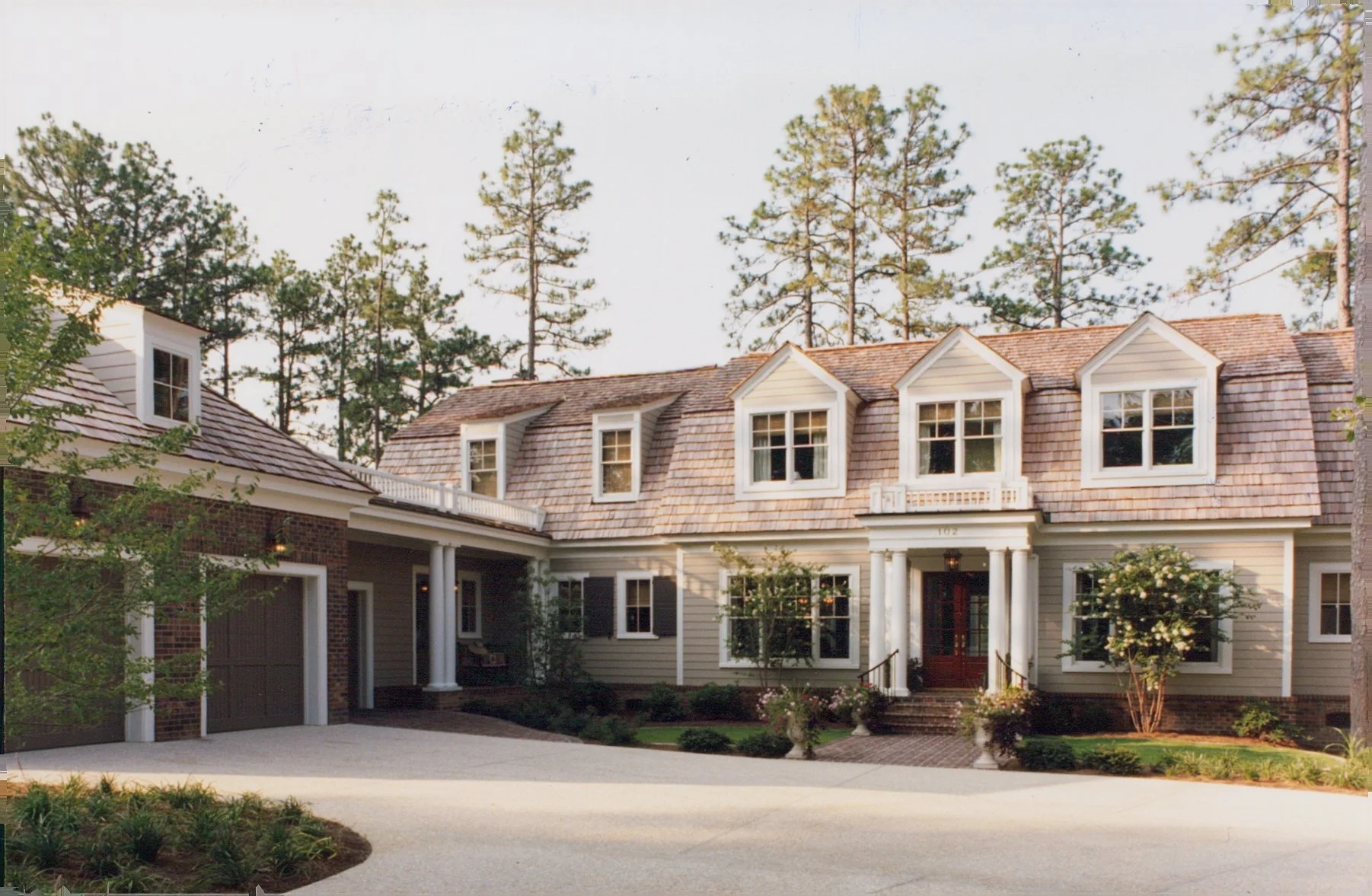 A large, two-story house with a wooden shingle roof, white siding, and multiple windows, surrounded by trees and a neatly landscaped yard with potted plants and a brick pathway leading to the front door.