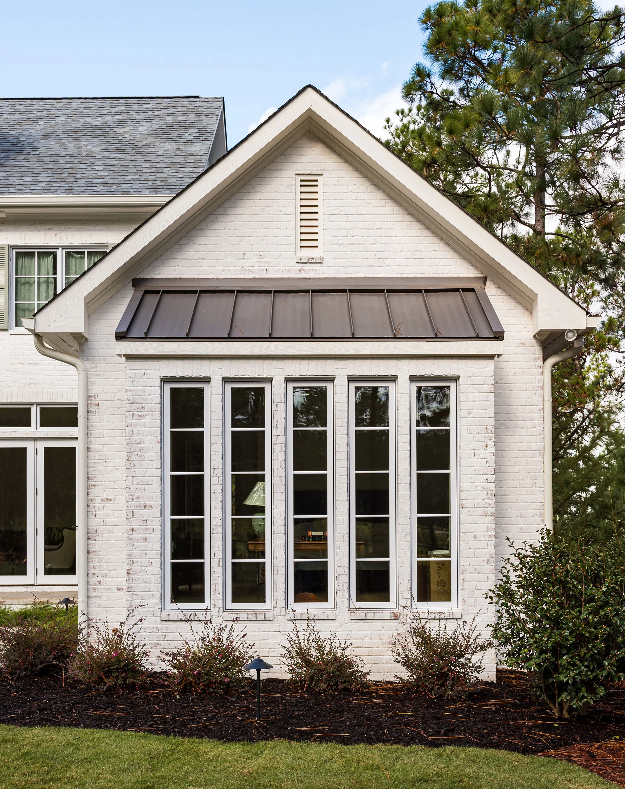 A white brick house with tall narrow windows, a dark metal awning, and a gable roof, with shrubs and a small garden in the front.