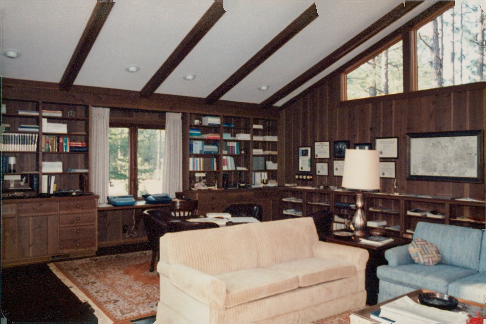 1980s Heritage Architectural design featuring a living room with wooden-paneled walls, large window, built-in bookshelves filled with books, and a seating area with a beige sofa, a blue armchair, and a side table with a lamp.