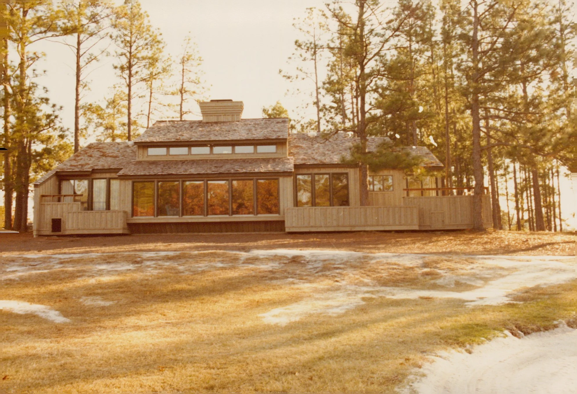 A large wooden house with multiple windows, situated on a slightly elevated ground surrounded by tall pine trees, during sunset.