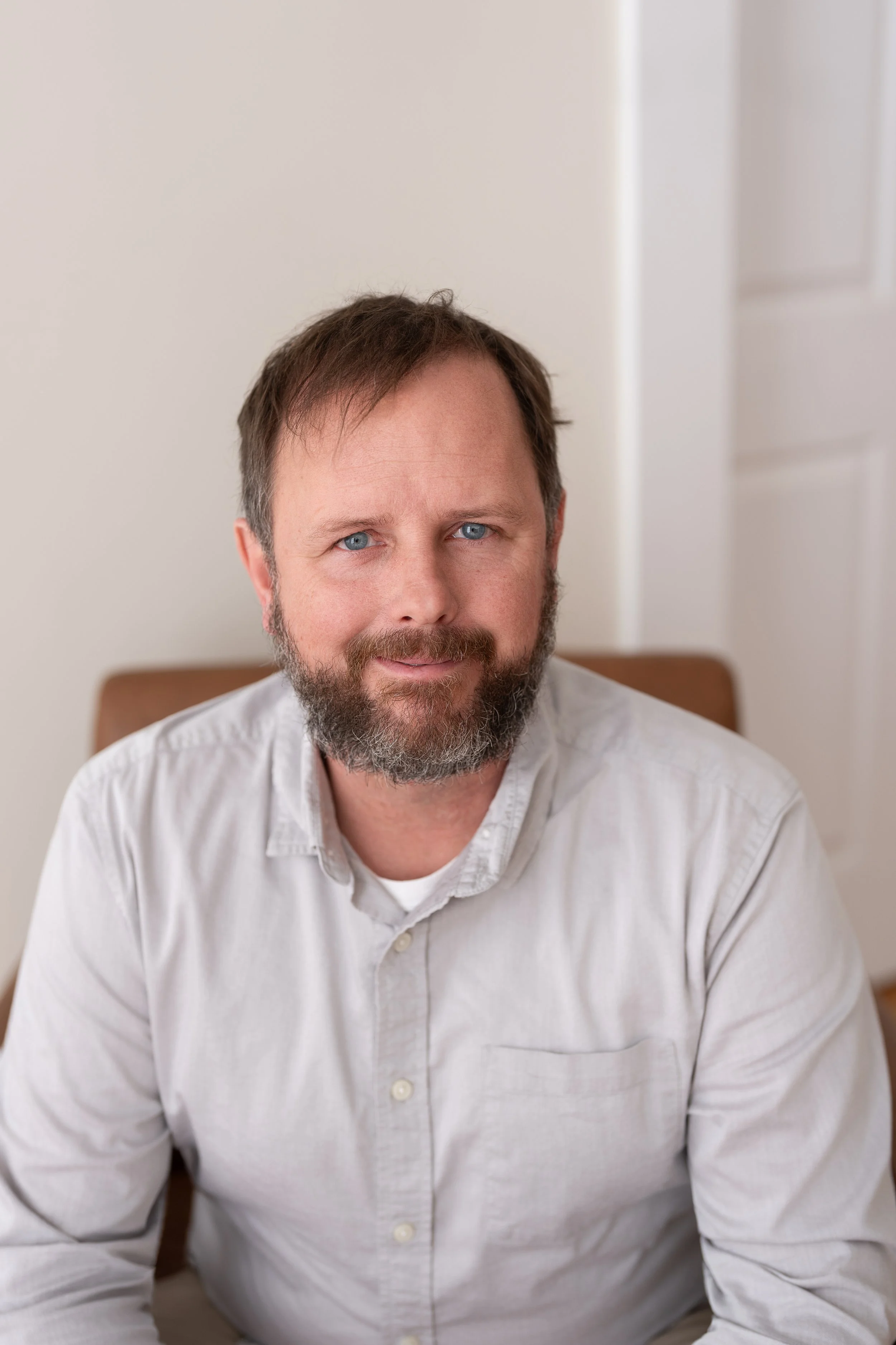 A man with light skin, blue eyes, and a beard seated indoors, wearing a light-colored button-up shirt, looking at the camera.
