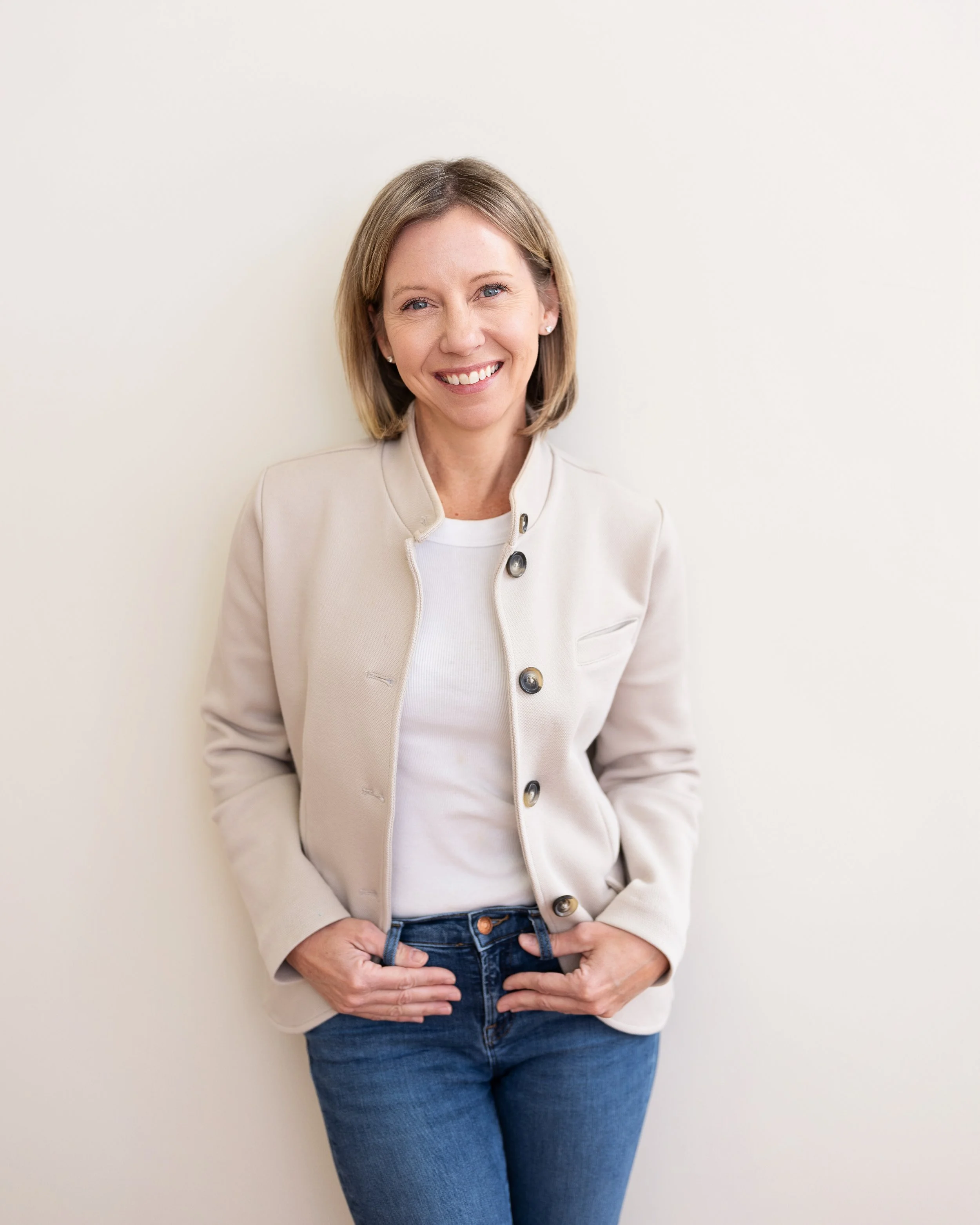 A smiling woman with shoulder-length blonde hair wearing a beige blazer, white shirt, and blue jeans, standing against a plain white wall.