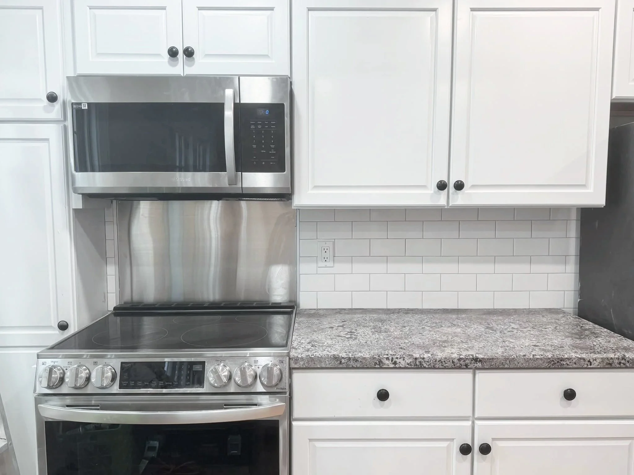 Kitchen with white cabinets, stainless steel microwave above a stainless steel electric stove, granite countertop, white subway tile backsplash, and electrical outlet.