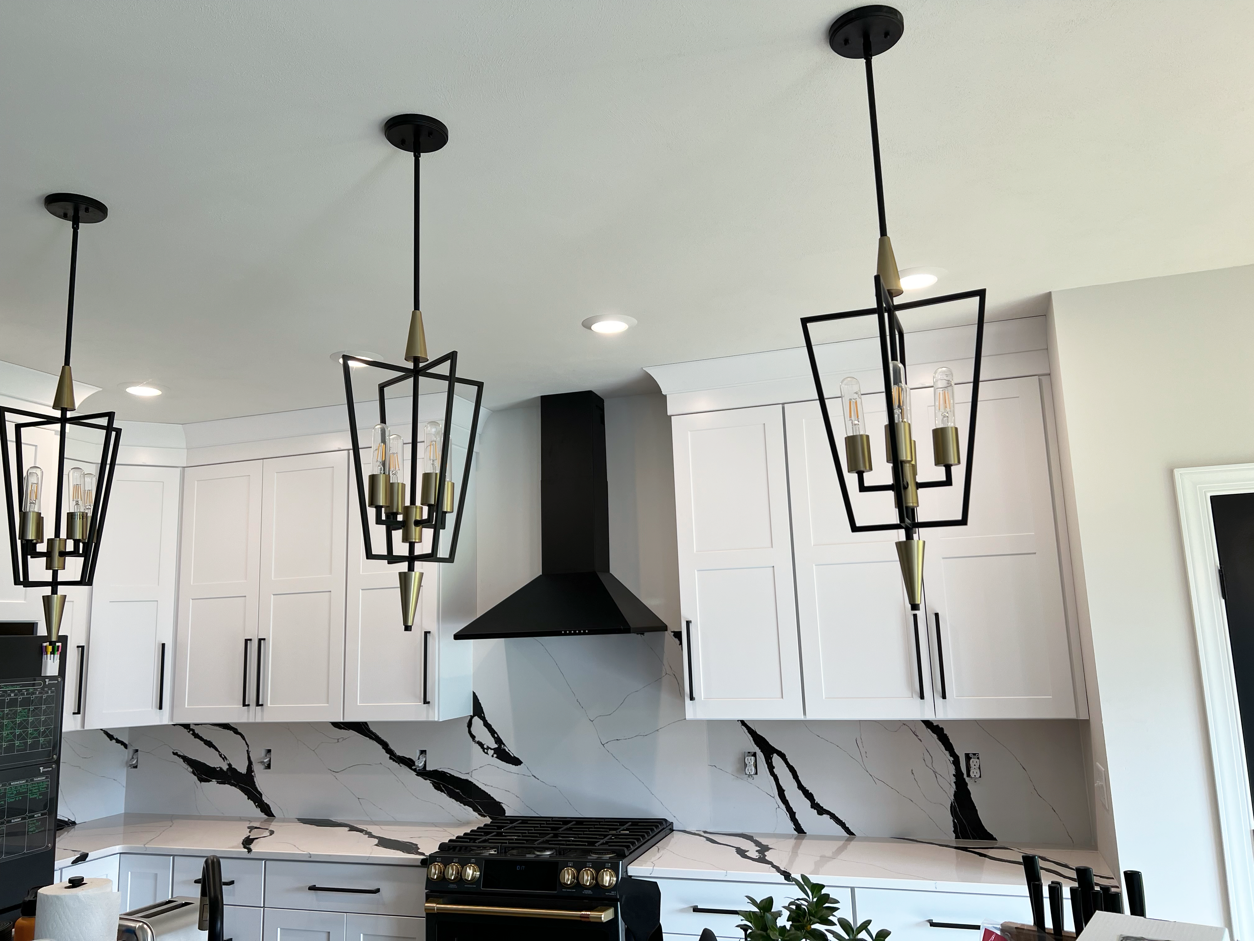 Modern kitchen with white cabinets, black hardware, marble backsplash, black range hood, and three black geometric pendant lights hanging from the ceiling.