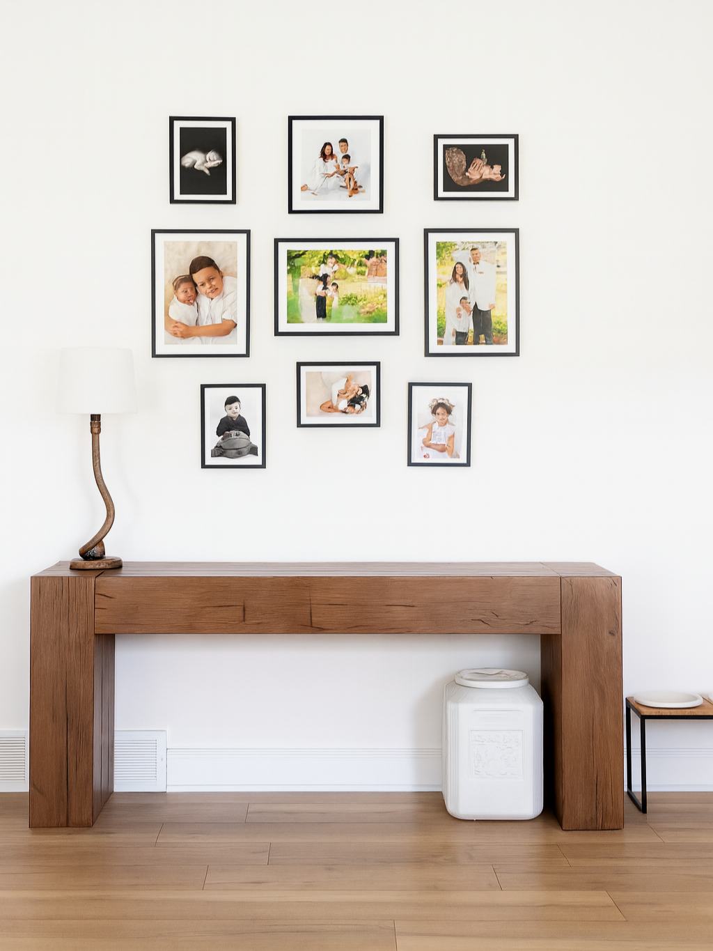 Wall with nine framed family and children photos arranged in a gallery style above a wooden console table, with a white lamp on the left, a white storage cube underneath, and a small table with a plate on the right.