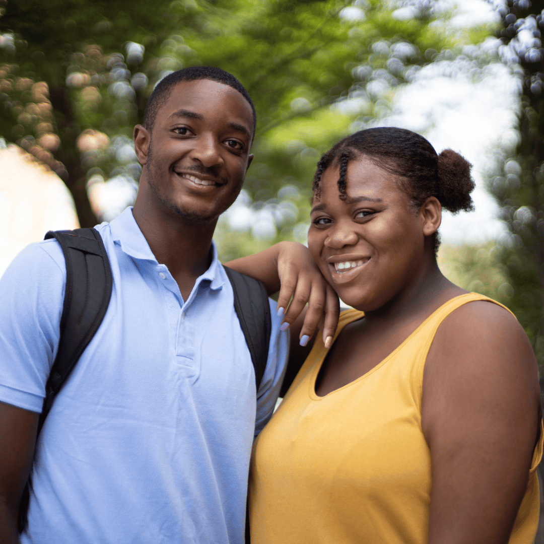 A young man and woman smiling outdoors, with the woman resting her hand on the man's shoulder, in front of a background of green trees.