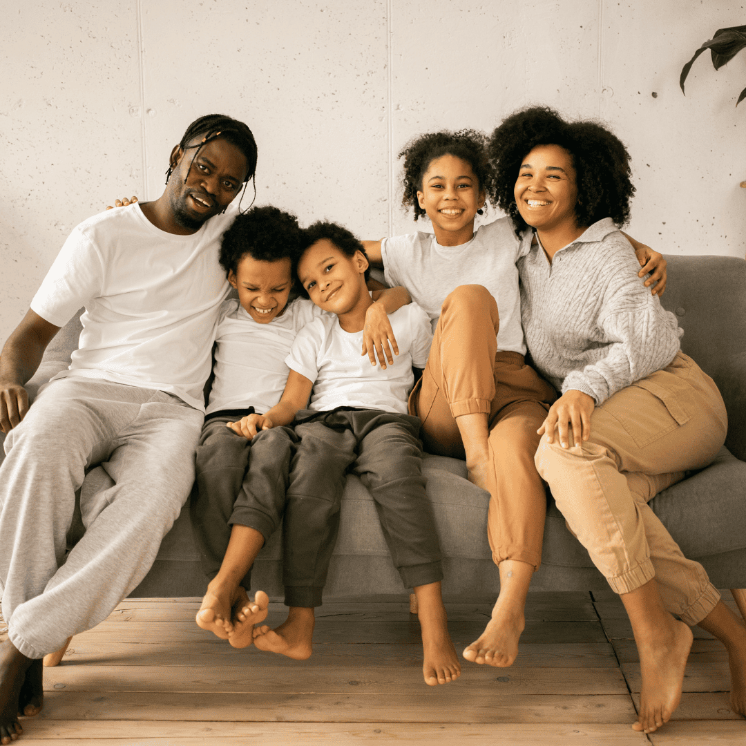 A family of six sitting on a gray couch, smiling and enjoying each other's company, in a cozy living room with a white wall background.