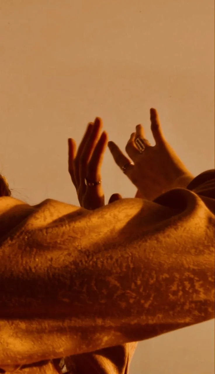 Close-up of a person's arm and hands with jewelry, raised against a warm-toned background.