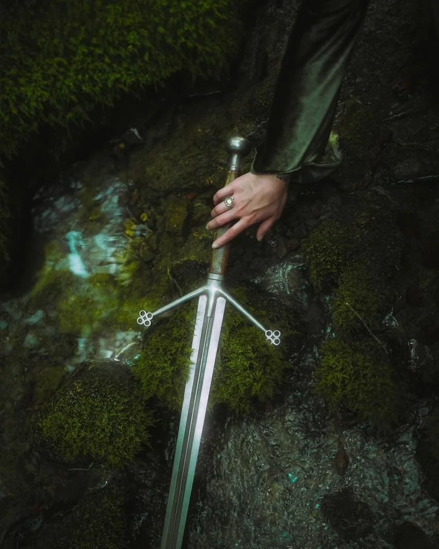 A person holding a sword with a mossy, rocky background and a small stream of water.