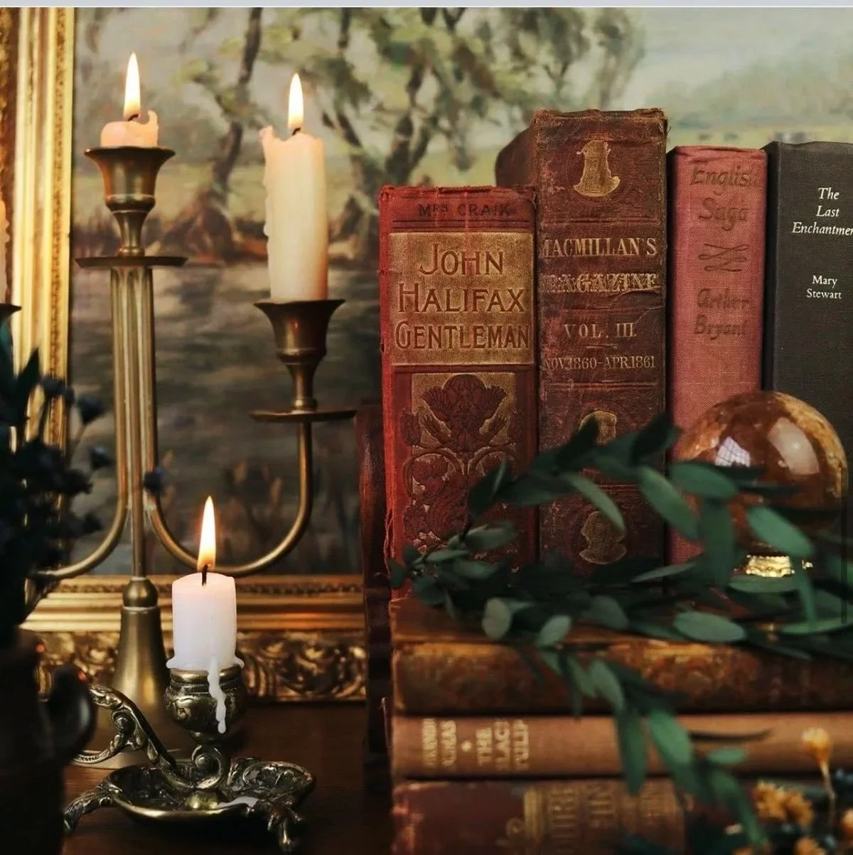 Stacked vintage books, a brass candelabra with lit candles, and a decorative glass orb on a wooden surface, with a landscape painting in the background.