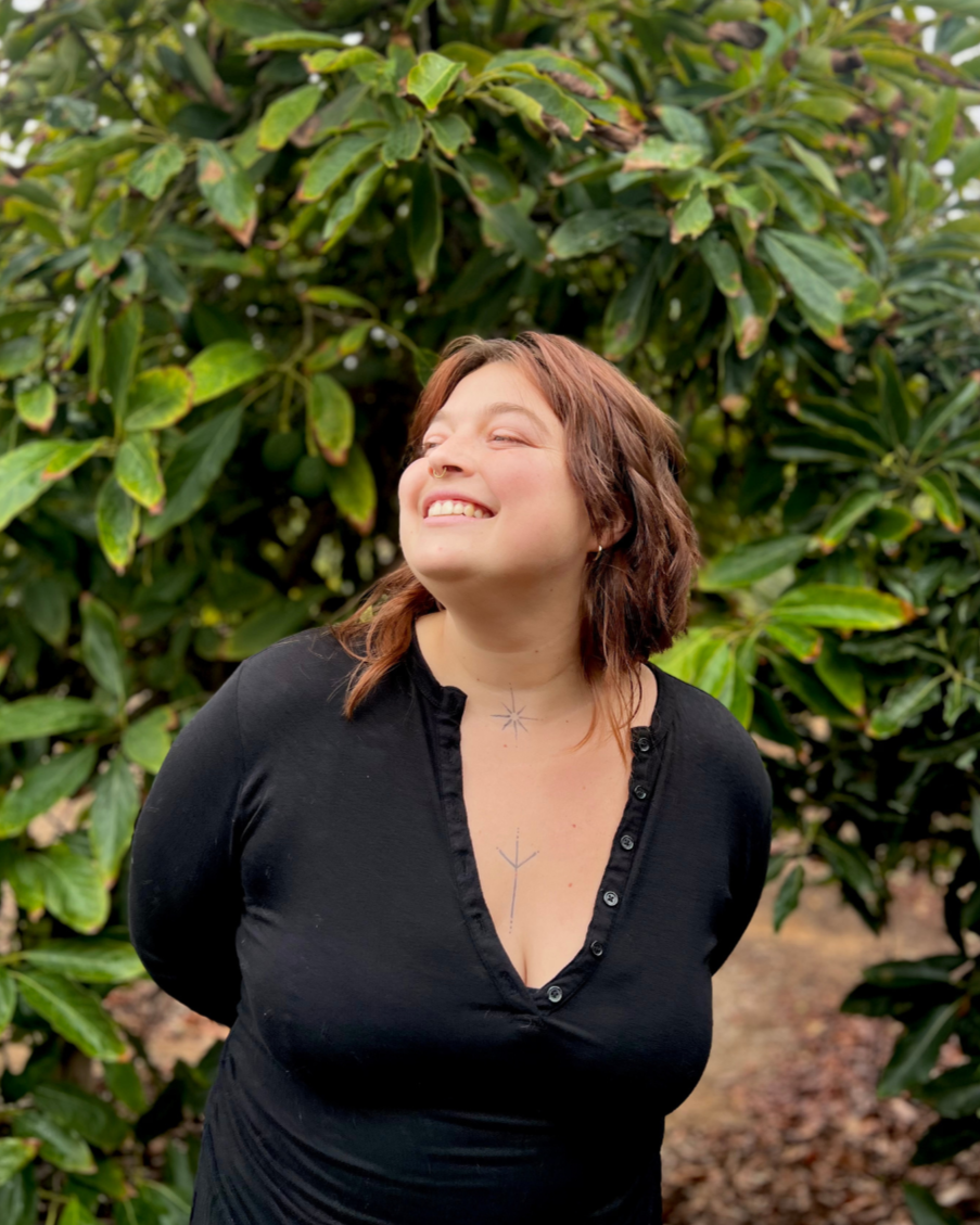 A smiling woman with brown hair standing outdoors in front of large green leafy bushes.
