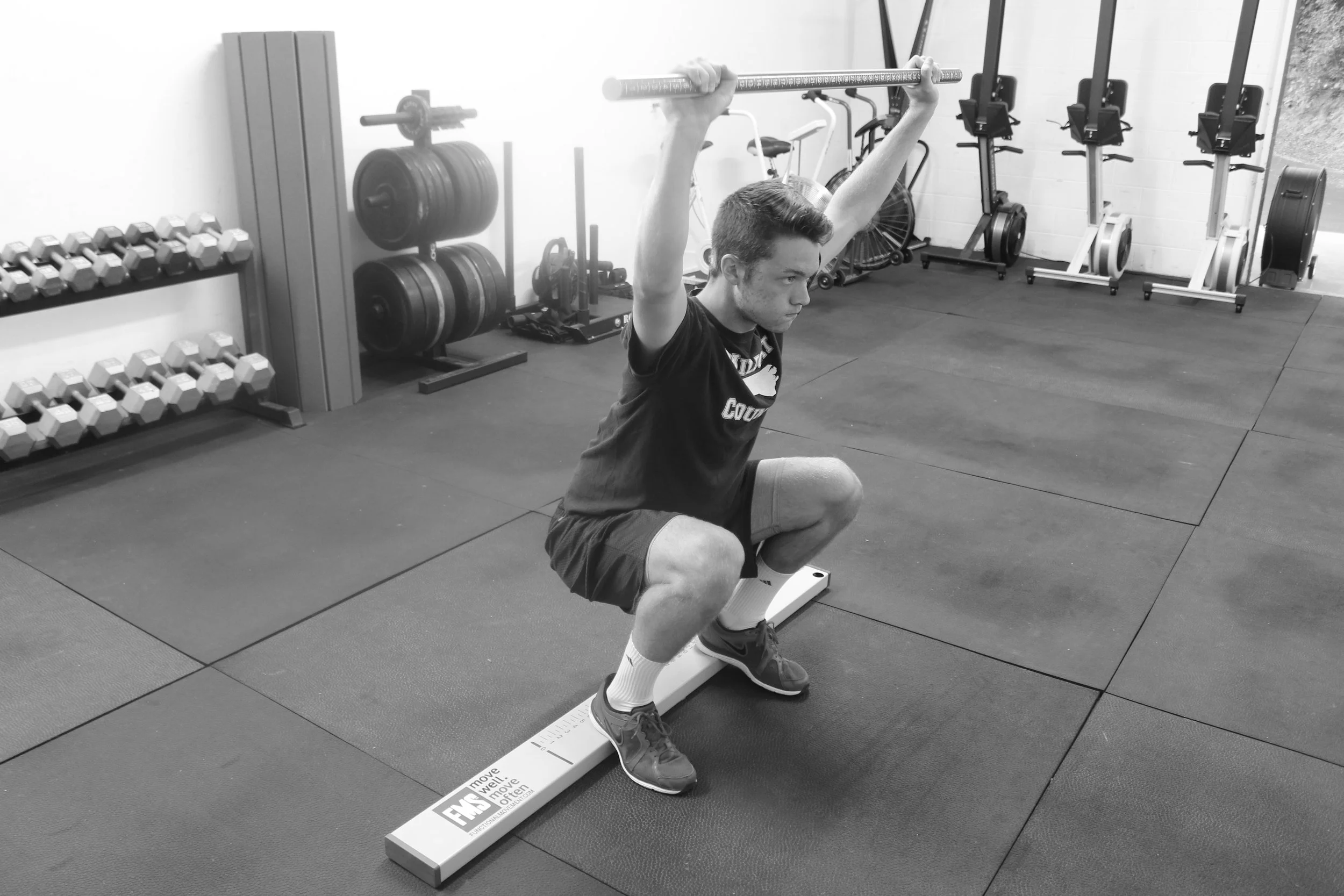 A young man in a gym squatting on a balance board while holding a barbell above his head.
