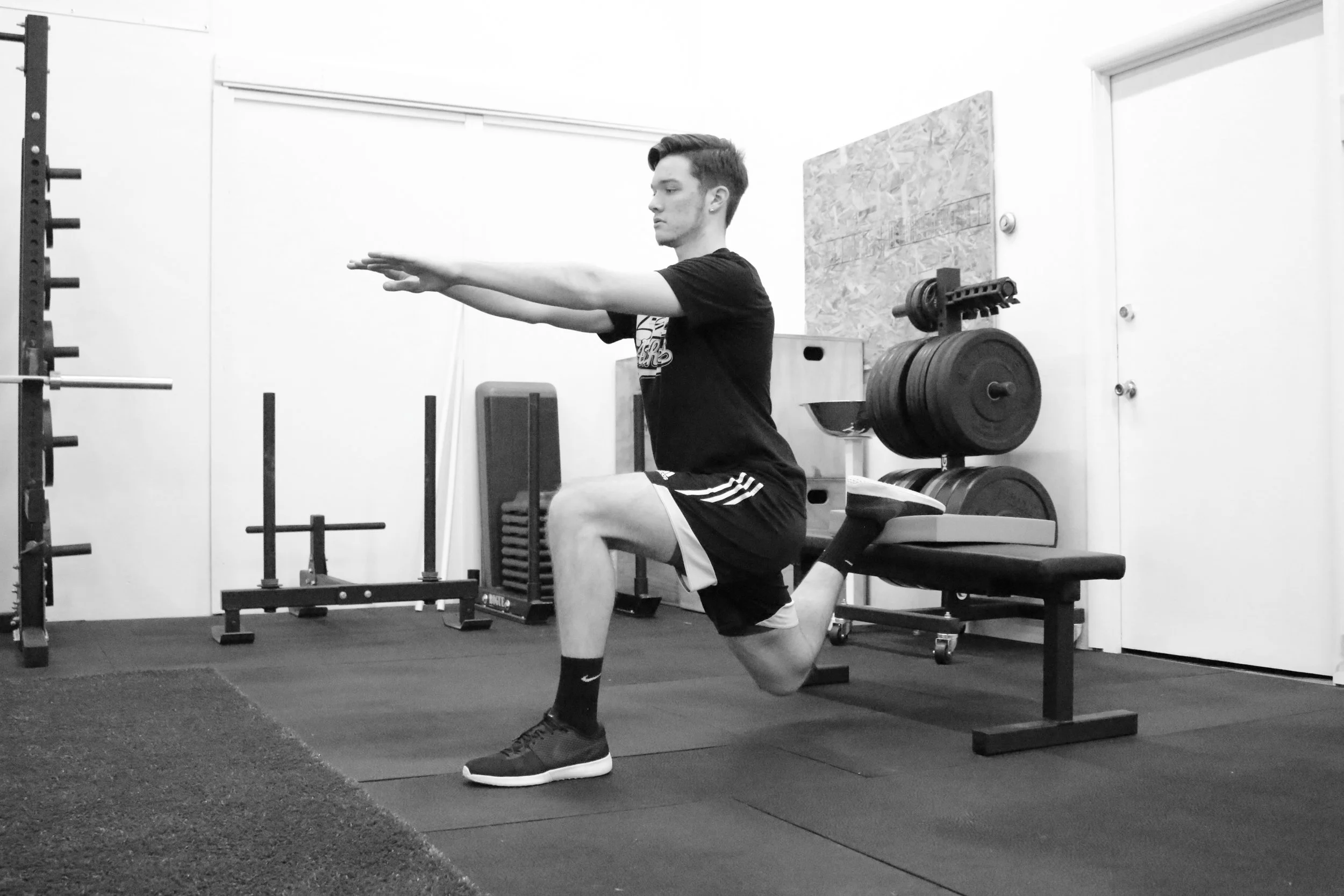 A young man in athletic wear is performing a lunge exercise on a bench in a gym, with weight plates and gym equipment visible in the background.