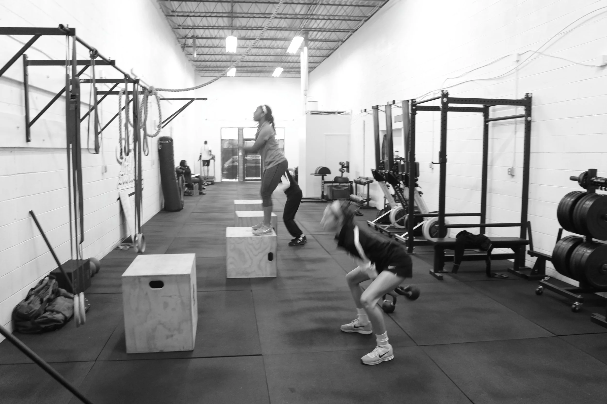 Two women exercising in a gym. One is jumping onto a wooden plyometric box, and the other is doing squats with a kettlebell. The gym has various workout equipment, including squat racks, weight plates, and resistance bands.